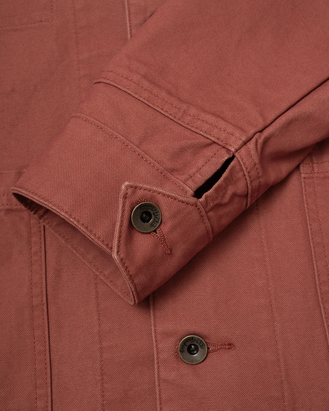 Close-up of the sleeve of an Imperfects - Cunningham Coat in Craft Canvas / Oxide, showing a terracotta-colored canvas fabric with a buttoned cuff and metal buttons.