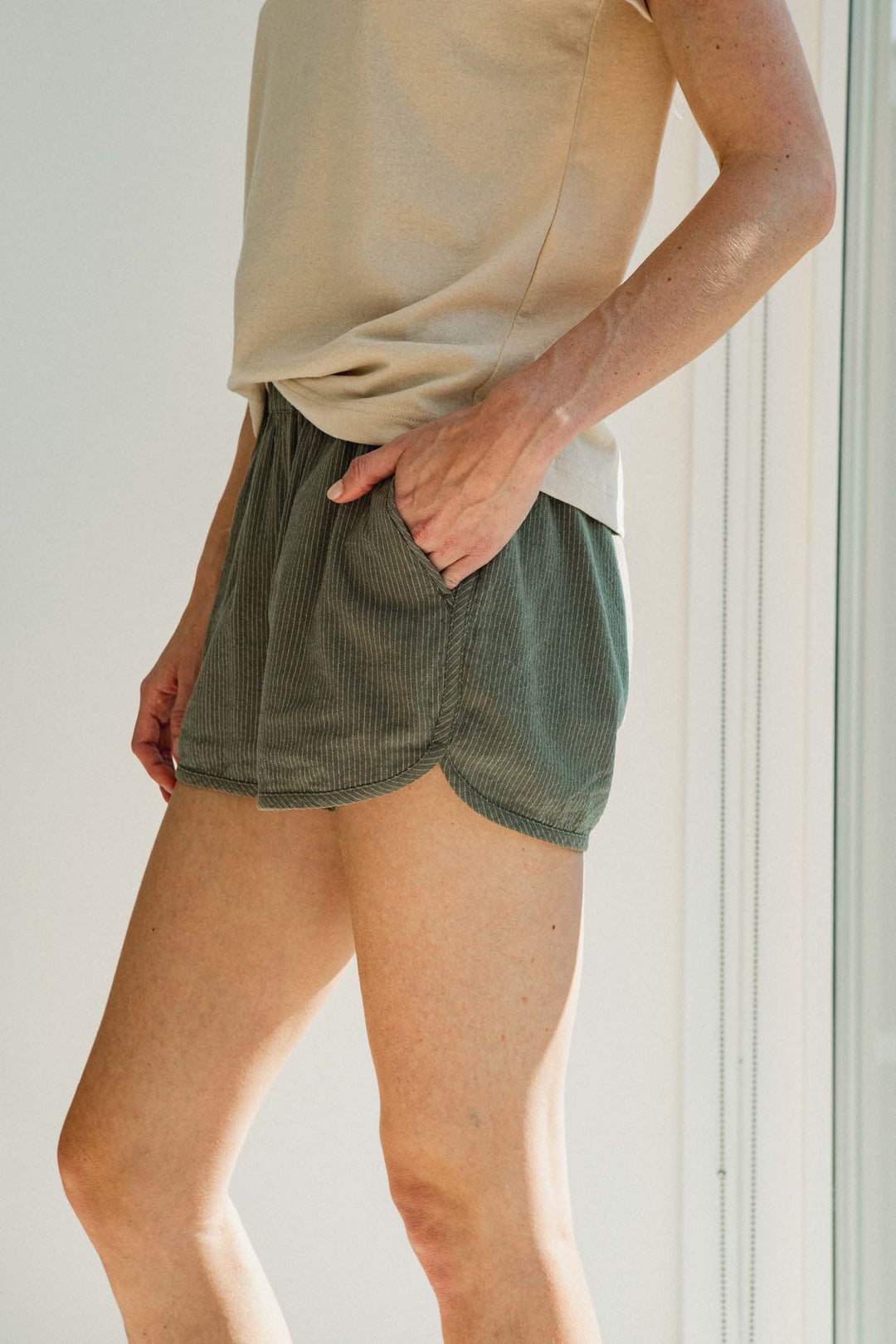 A person wearing Bridge & Burn Luca Track Short / Olive Pinstripe linen blend track shorts with a curved hem and side pockets.