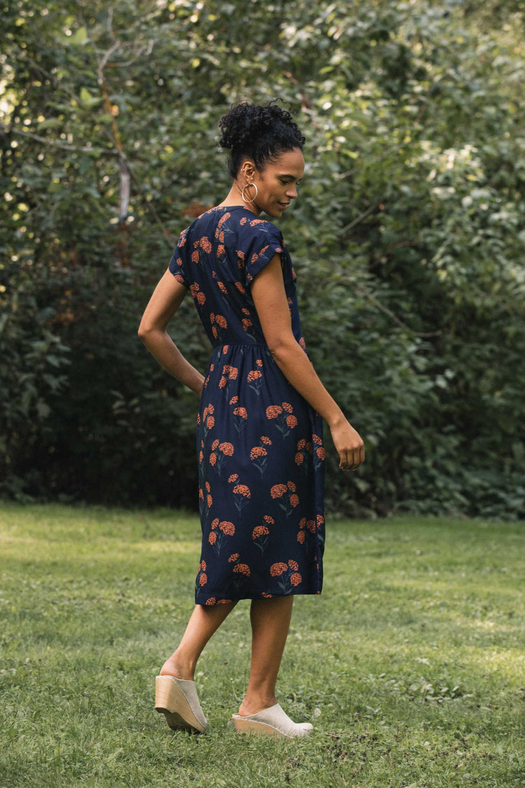 A person stands in a park wearing a Bridge & Burn Ira Dress / Wildflower in navy blue with a repeating orange wildflower print, paired with light-colored mules.