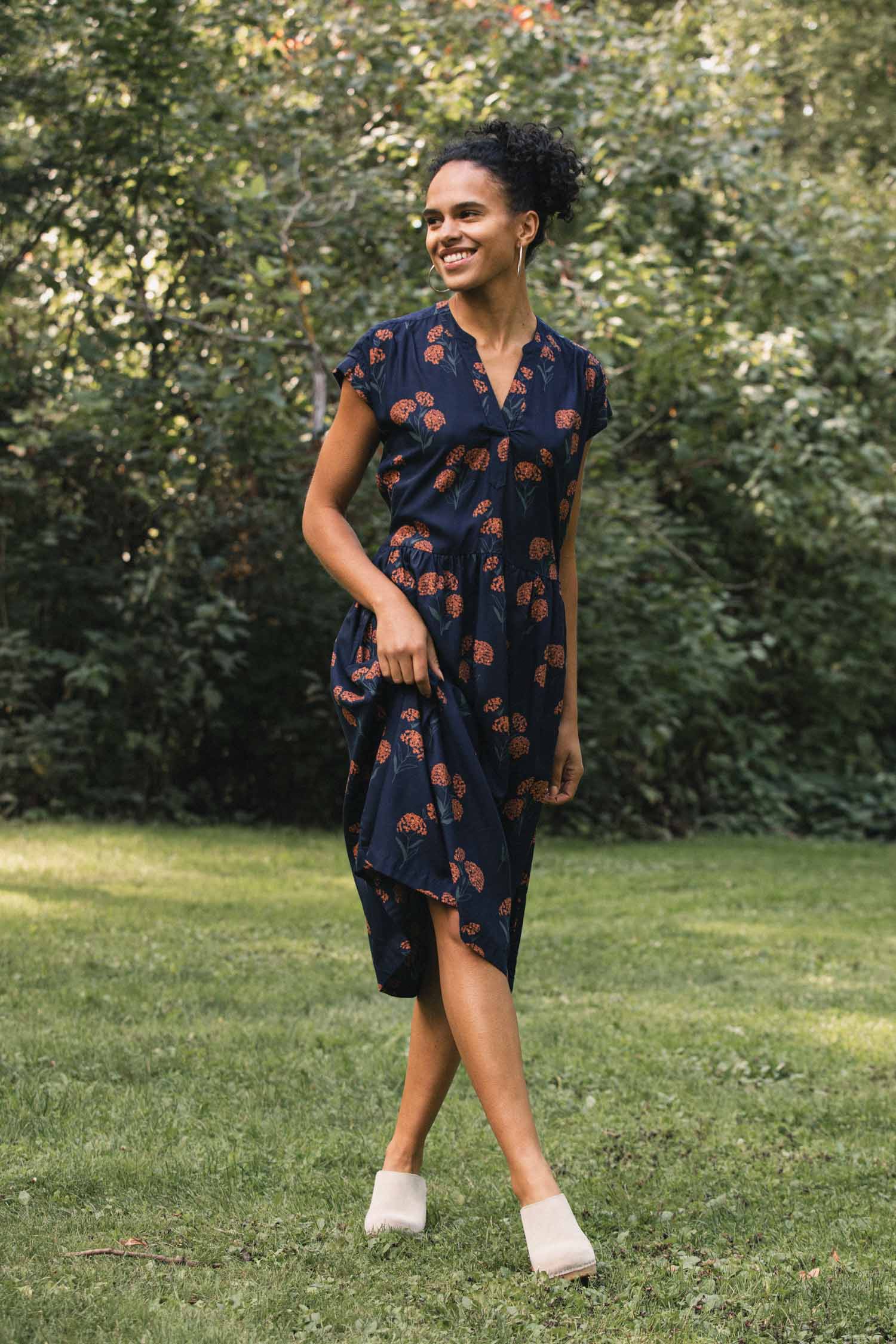 A person wearing a wrinkle-resistant, navy blue Ira Dress / Wildflower midi dress with a repeating orange wildflower print and light-colored clogs, standing in a park.