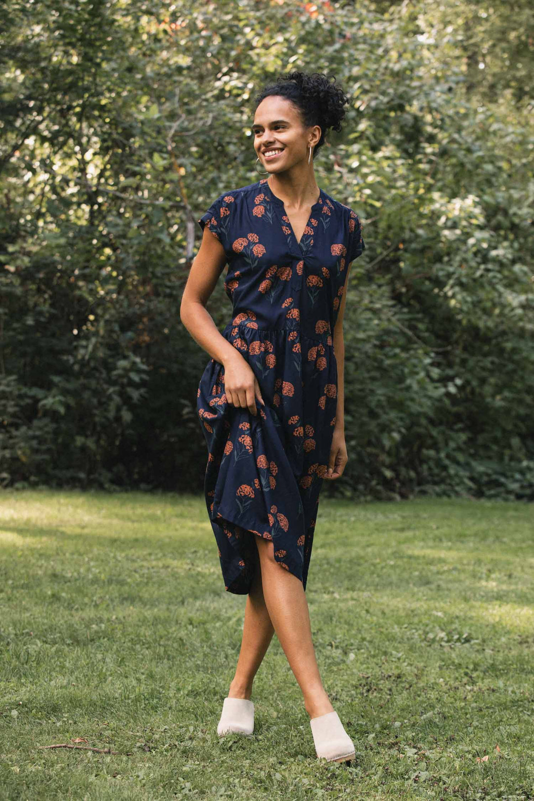 A person wearing a wrinkle-resistant, navy blue Ira Dress / Wildflower midi dress with a repeating orange wildflower print and light-colored clogs, standing in a park.