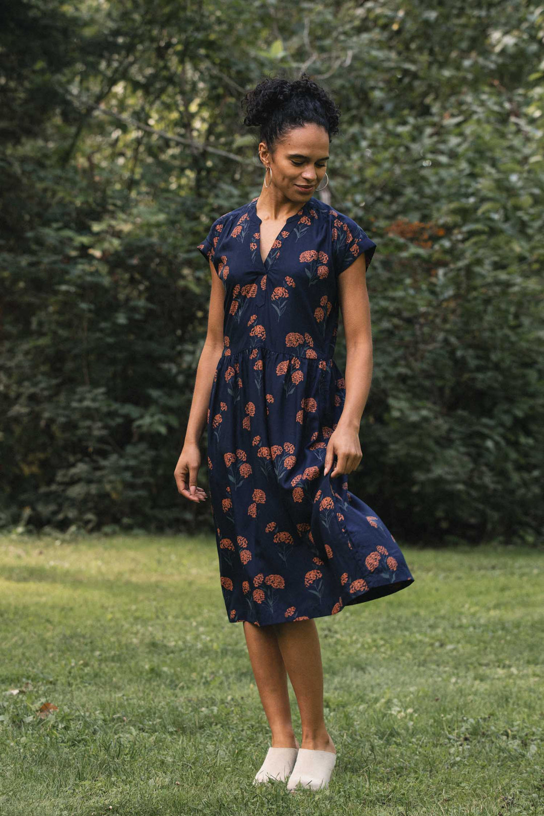 A person stands on a lawn wearing a Bridge & Burn Ira Dress in Wildflower, a sustainable navy blue midi pullover dress with a repeating rust-colored wildflower print and cap sleeves.