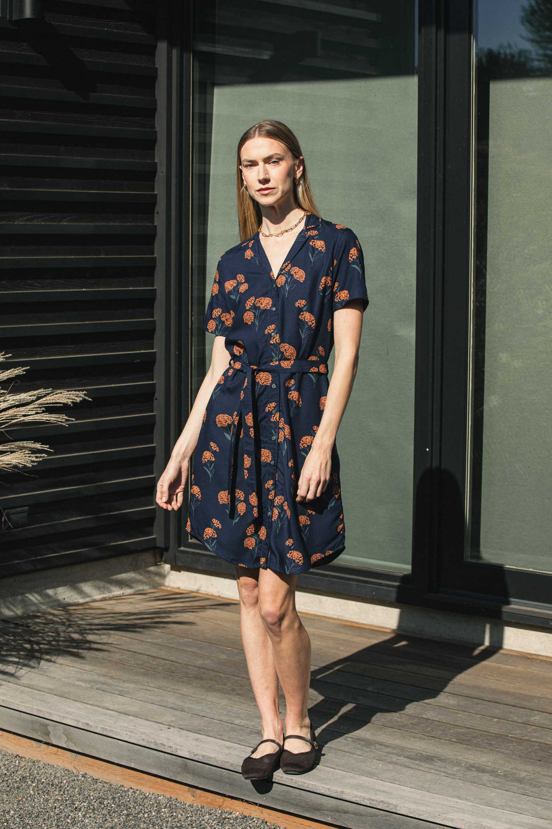 A woman wears a Bridge & Burn Sofia Shirt Dress / Wildflower, a sustainable, navy blue modal dress with a self-belted waist and an orange wildflower print.