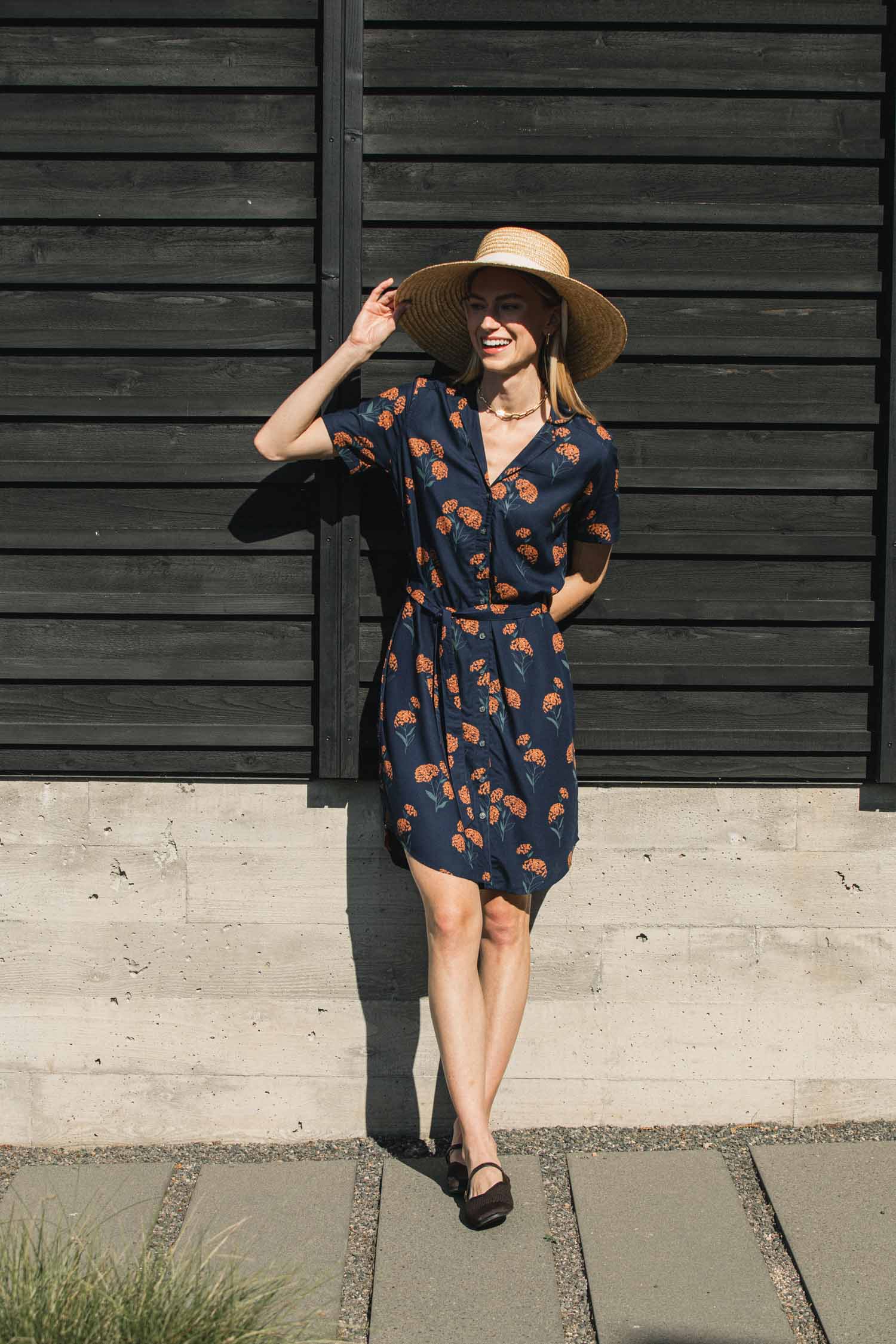 A woman wears a straw hat and a navy blue, short-sleeved Sofia Shirt Dress / Wildflower featuring a small orange floral print.