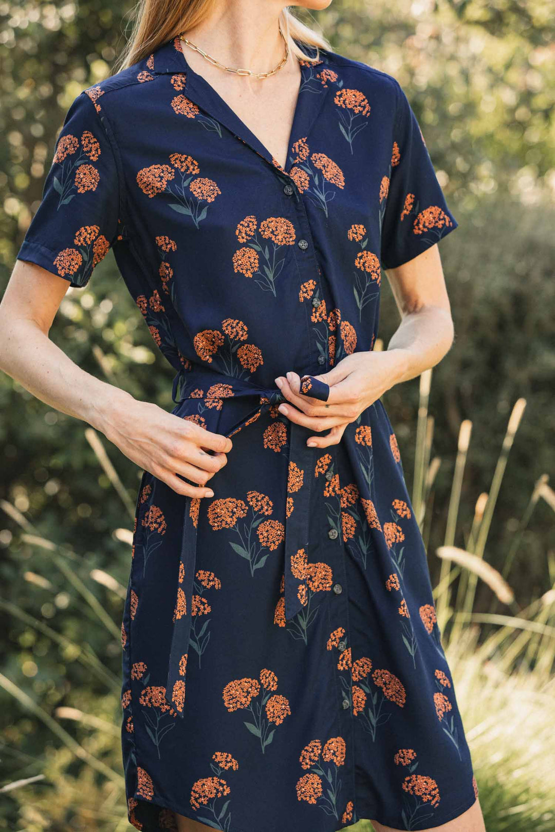 A person wearing the Bridge & Burn Sofia Shirt Dress / Wildflower, a sustainable, navy blue modal button-down dress featuring an orange wildflower print and a matching tie waist.