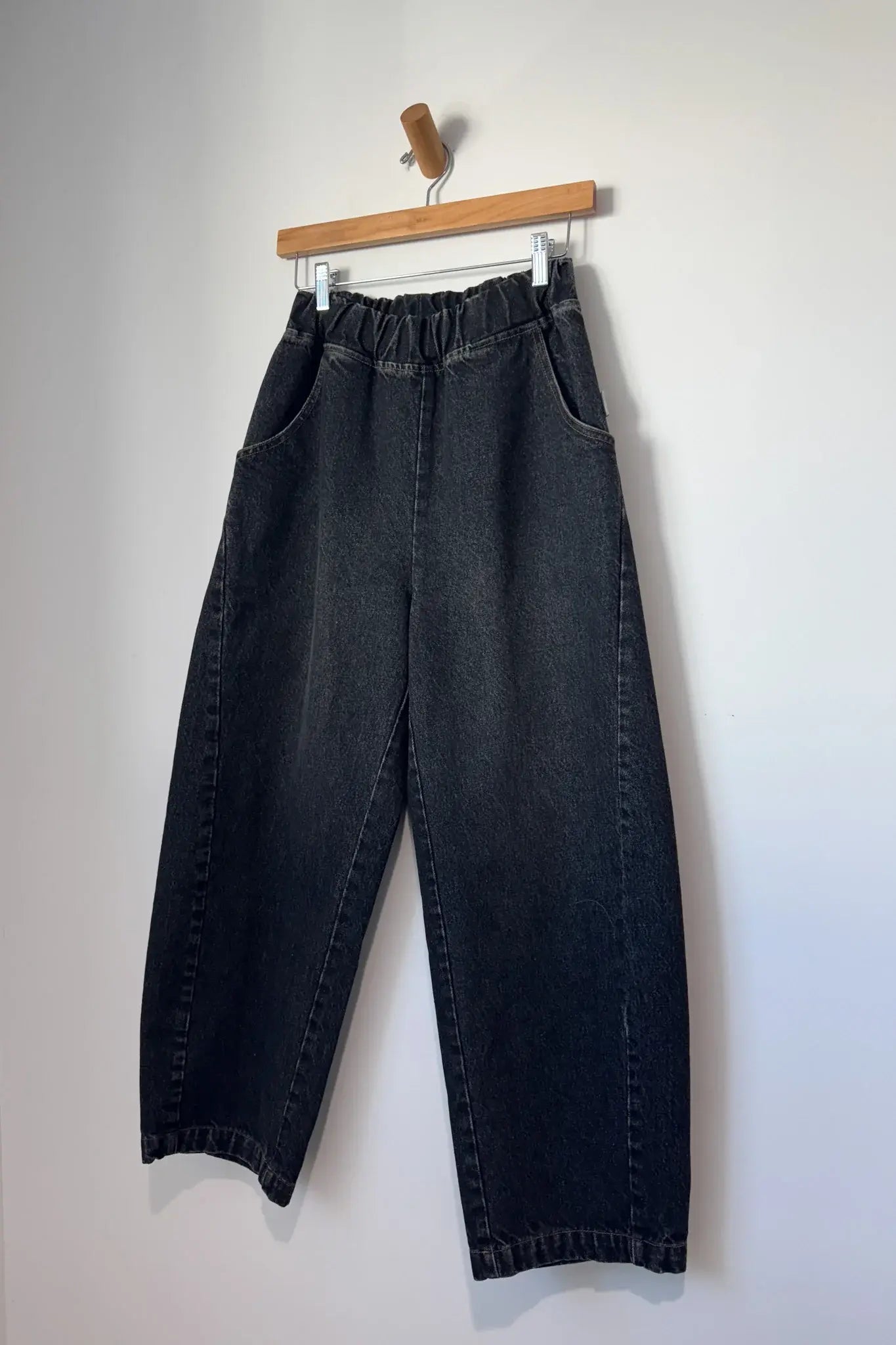 Le Bon Shoppe - Arc Pant in black denim, featuring a high-waisted fit, elastic waistband, and curved side pockets, hanging on a wooden hanger against a white wall.