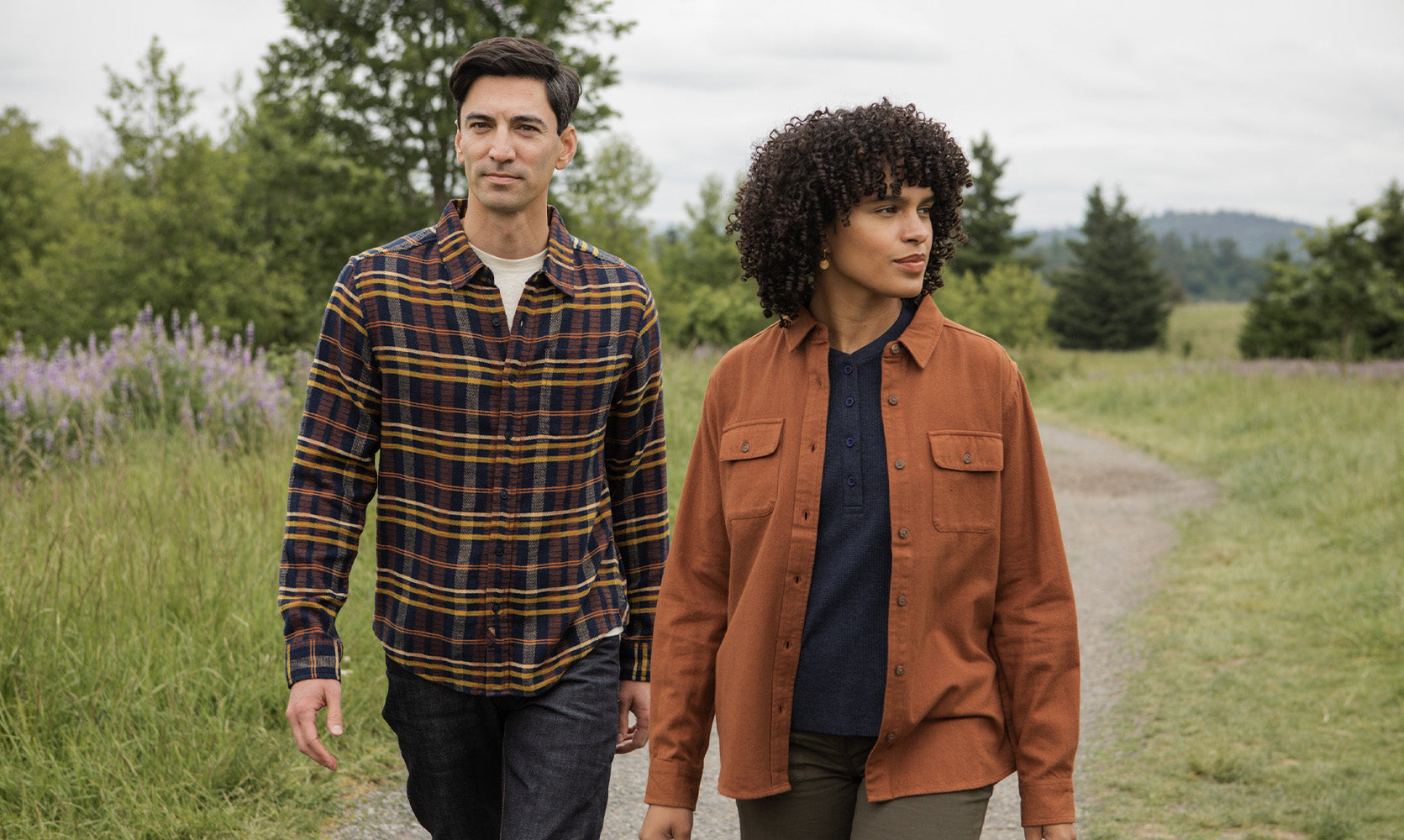 A man and a woman walk together on a gravel path surrounded by grass and trees. The man wears a plaid shirt and jeans; the woman wears a brown jacket and dark pants. Both appear relaxed and outdoors.