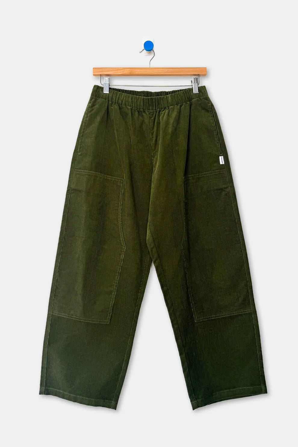 Le Bon Shoppe Gardener’s Pant in Bay Leaf corduroy, featuring an elastic waist and large patch pockets on the thighs, hanging on a wooden hanger.