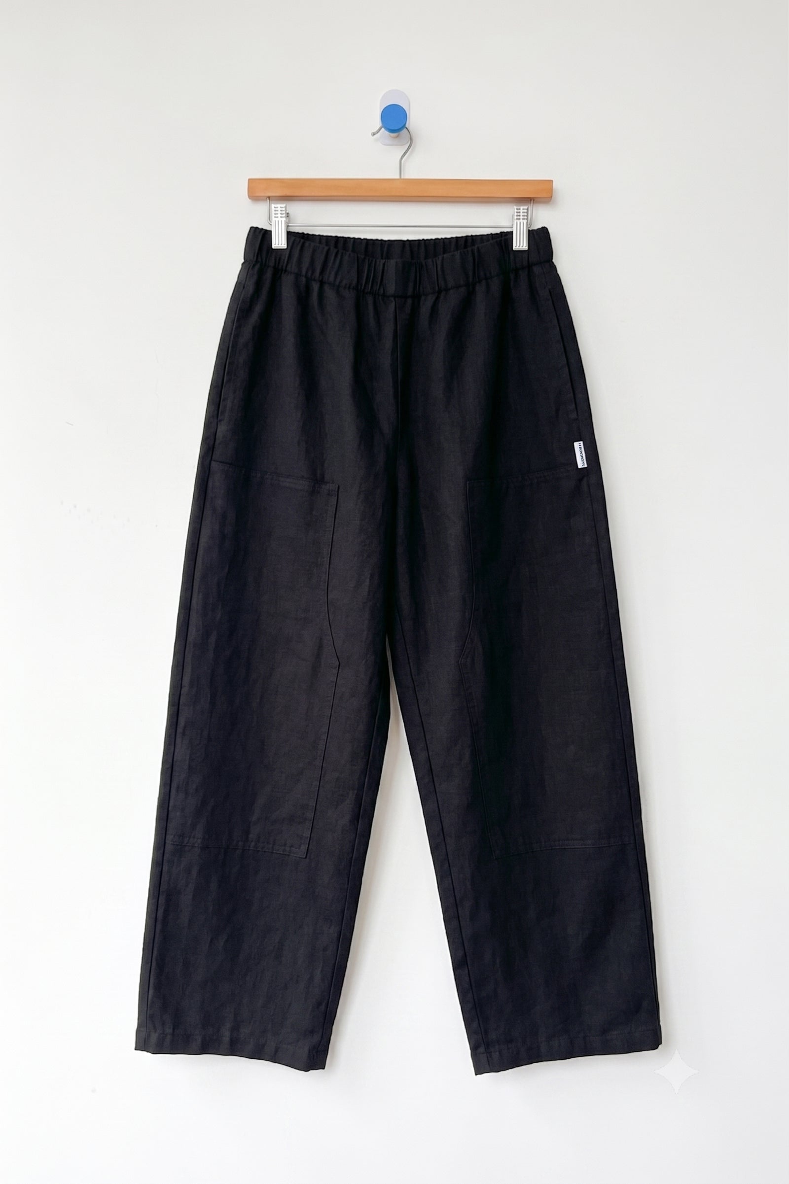 A pair of Le Bon Shoppe - Gardener's Pant in black, featuring a relaxed silhouette with an elastic waistband and spacious front pockets, crafted from lightweight cotton-nylon fabric, displayed on a wooden hanger against a plain white wall.