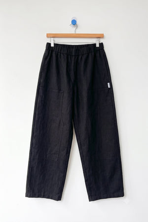 A pair of Le Bon Shoppe - Gardener's Pant in black, featuring a relaxed silhouette with an elastic waistband and spacious front pockets, crafted from lightweight cotton-nylon fabric, displayed on a wooden hanger against a plain white wall.