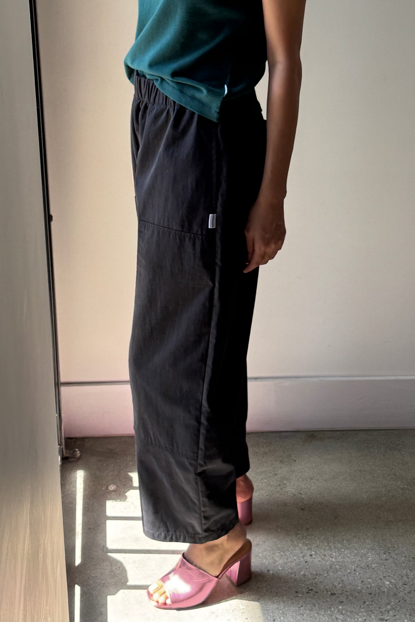 A person stands indoors in sunlight, wearing a green top and Le Bon Shoppe's Gardener's Pant in Black—crafted from lightweight cotton-nylon for a relaxed silhouette. Shiny pink open-toe heeled sandals complete the look, with only their torso and legs visible.