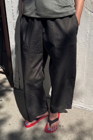 A person stands on the sidewalk with hands in their pockets, wearing Le Bon Shoppe - Gardener's Pant in Black from Le Bon Shoppe, which features a relaxed silhouette. They pair it with a gray shirt, black flip-flops with bright red soles, and blue nail polish. Sunlight casts shadows on the ground around them.