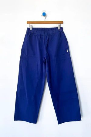 The Le Bon Shoppe Gardener's Pant in Deep Blue, featuring a relaxed silhouette and elastic waistband, hangs on a wooden hanger against a white background.