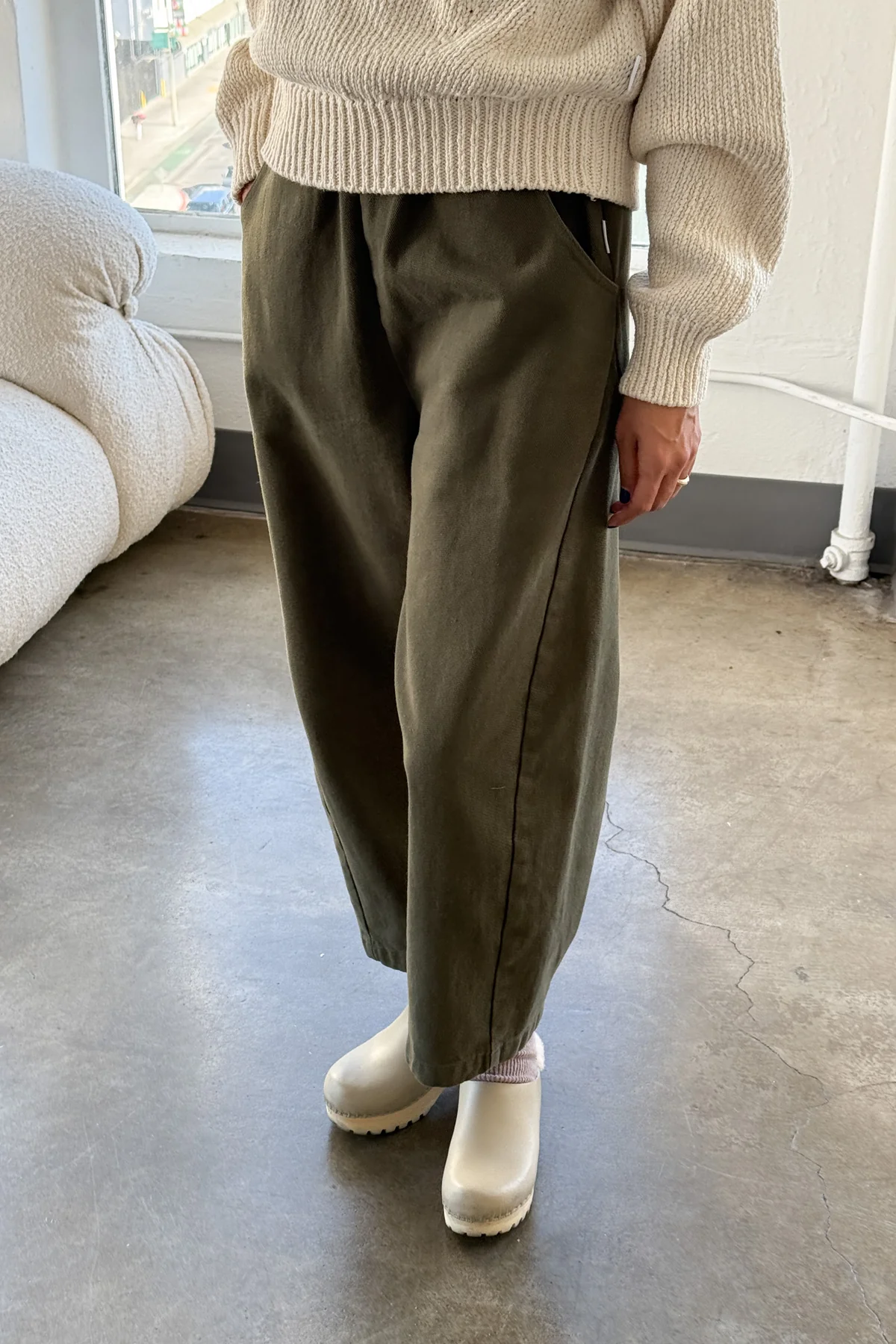 Only the lower body is visible: standing indoors on concrete, wearing Le Bon Shoppe - Arc Pant / Olive high waist wide-leg pants, a cream knit sweater, and white slip-on clogs.