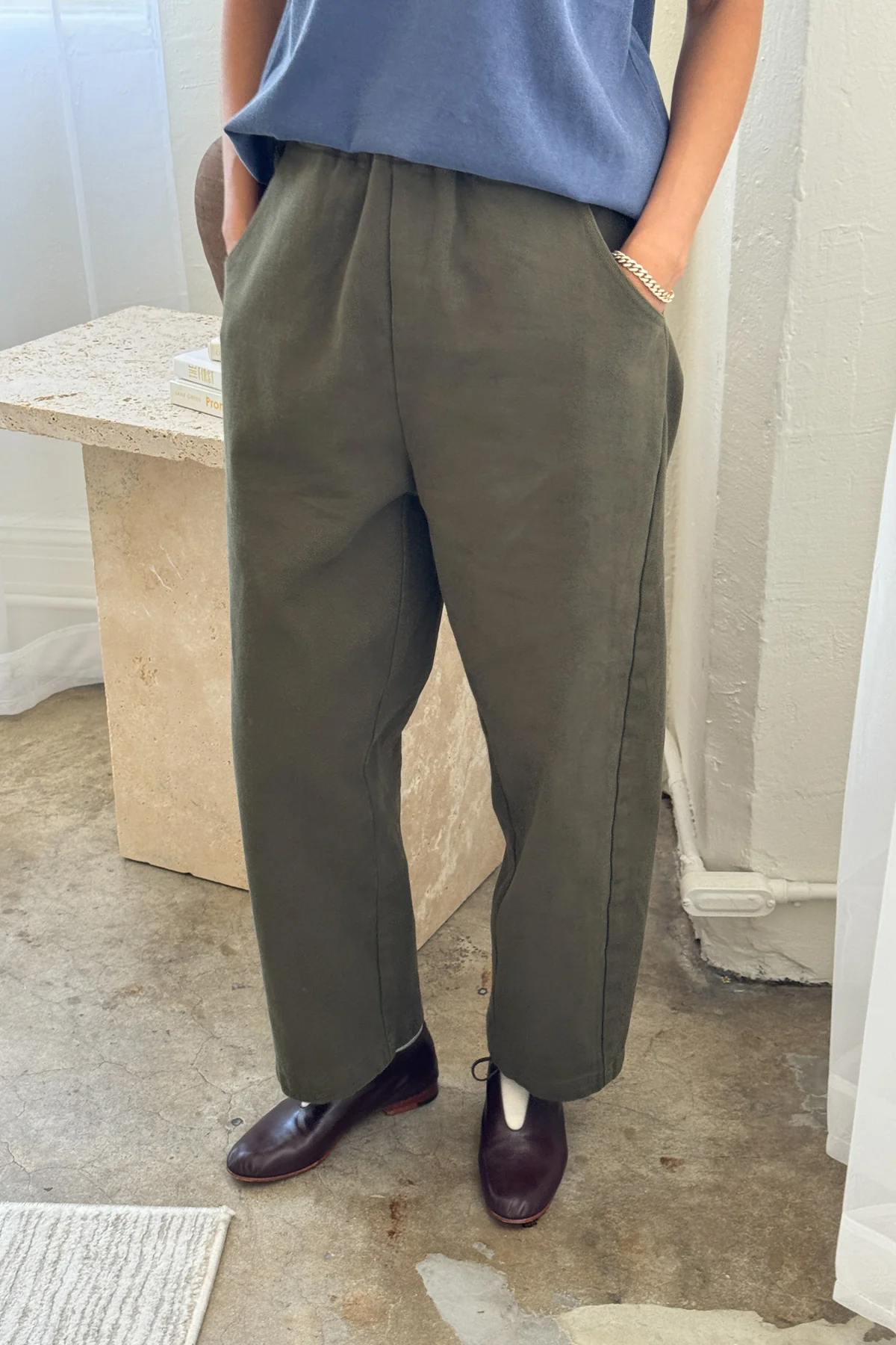 A person stands indoors with hands in pockets, wearing Le Bon Shoppe - Arc Pant in Olive and a blue top, paired with dark brown shoes. A stone table and textured concrete floor are visible; the person's face is not shown.