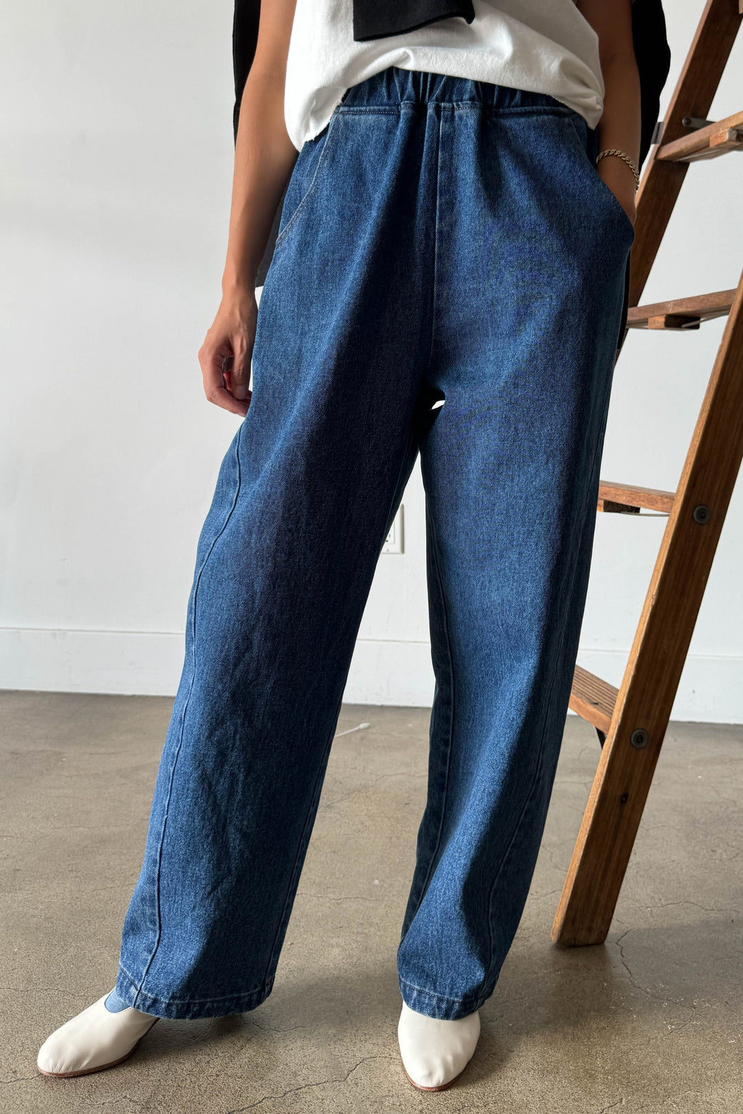 A person stands indoors on a concrete floor near a wooden ladder, wearing the Le Bon Shoppe - Long Arc Pant / Blue Denim by Le Bon Shoppe—loose-fitting blue jeans crafted from heavyweight denim—paired with a white shirt and white pointed shoes. The upper part of their face is not visible.