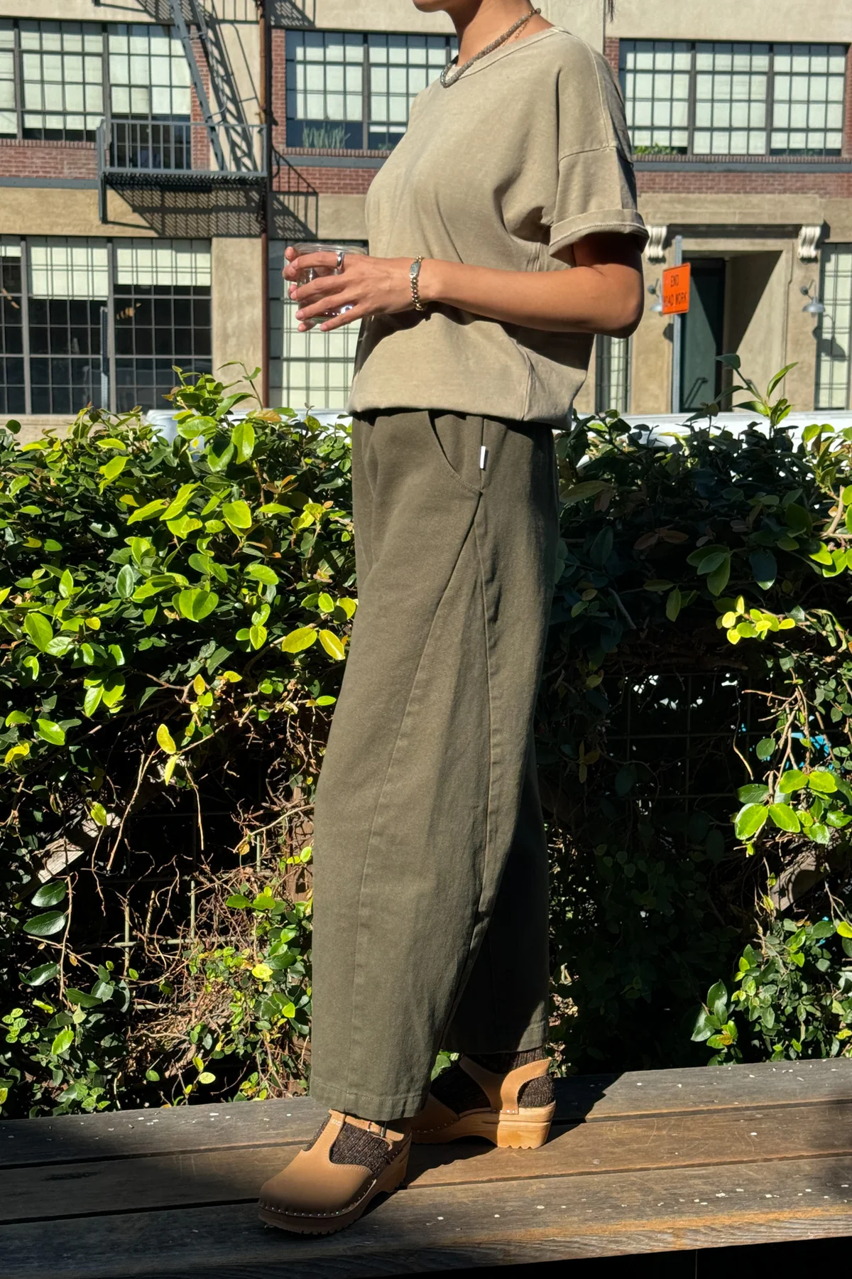 A person stands outdoors in sunlight holding a drink, wearing Le Bon Shoppe's Arc Pant in Olive with a matching oversized tee and tan clogs. Patterned socks, green bushes, and a building with large windows are in the background.