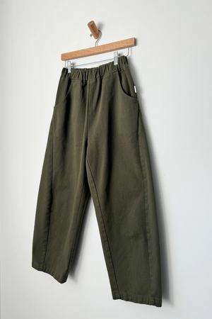 The Le Bon Shoppe Arc Pant in Olive features a high waist, wide-leg canvas design with an elastic waistband and side pockets, shown hanging on a wooden hanger against a plain white wall.