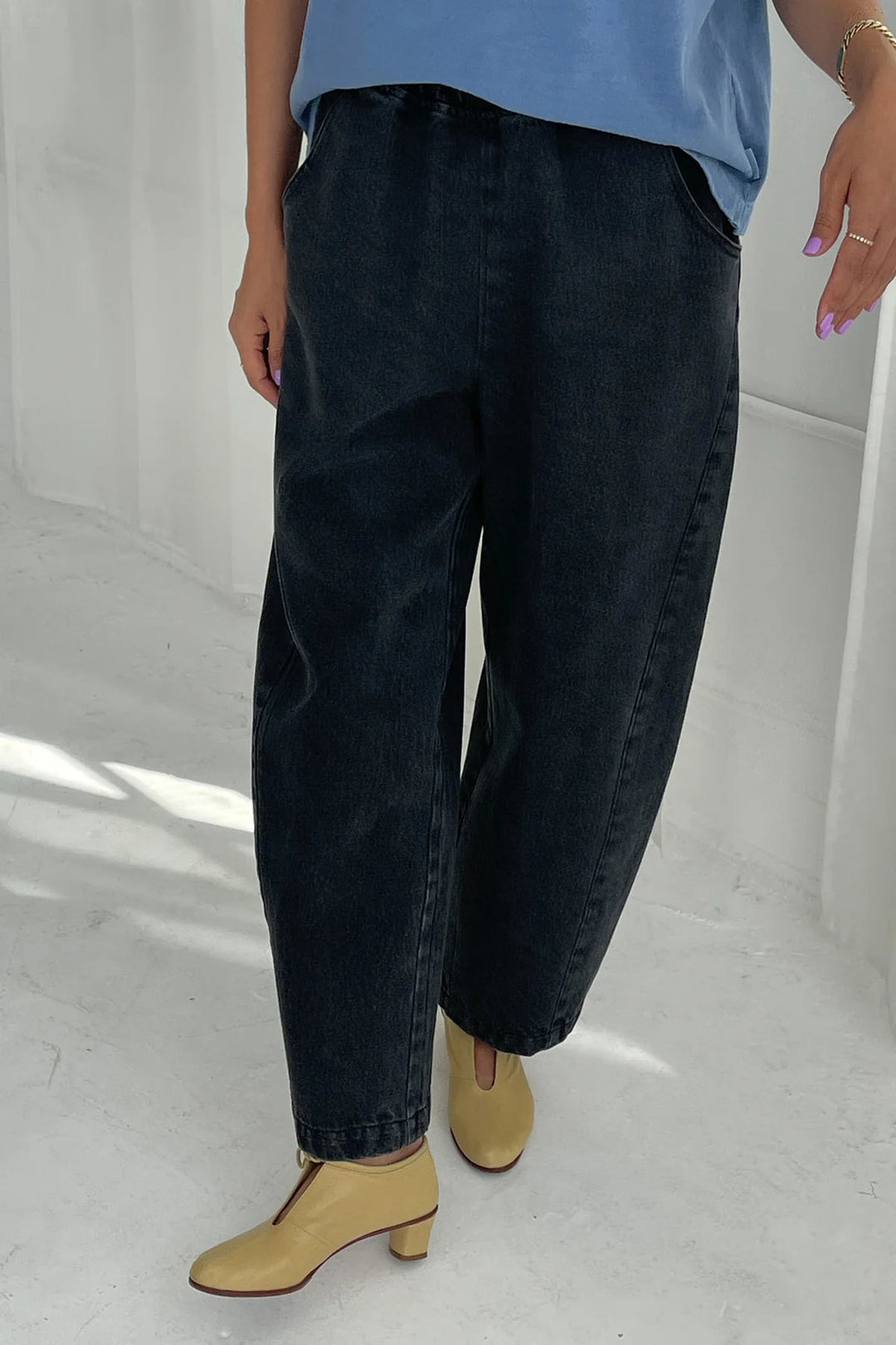 A person is shown from the waist down wearing the Le Bon Shoppe - Arc Pant in Black Denim by Le Bon Shoppe, paired with a light blue top and beige heeled shoes, standing on a white floor.