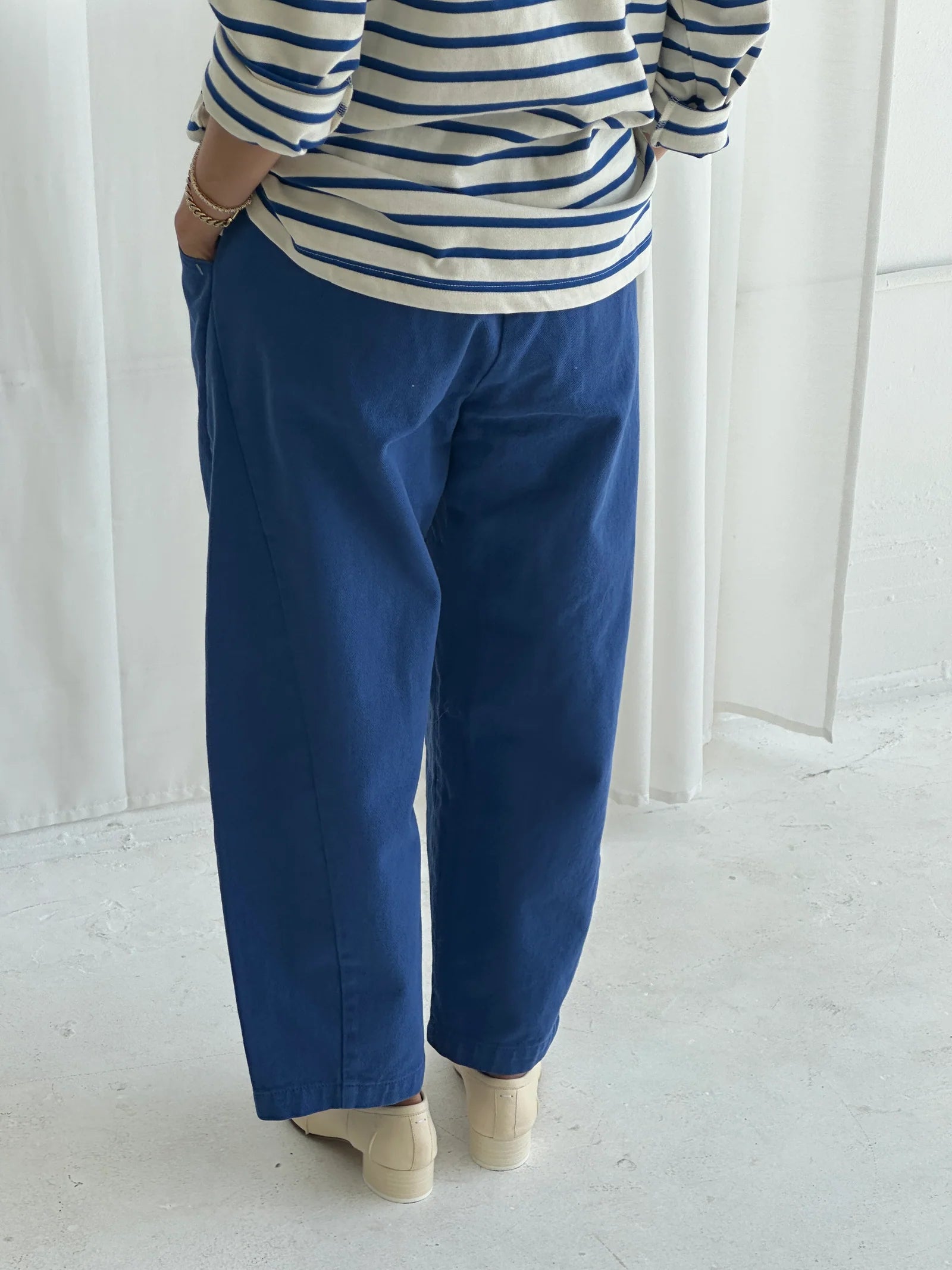 A person stands on a light floor, wearing Le Bon Shoppe - Arc Pant / Azure high-waist loose blue denim, paired with a white and navy striped long-sleeve shirt and white shoes. Only their lower body and part of their arms are visible.