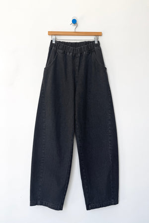 A pair of Le Bon Shoppe - Long Arc Pant in black denim, featuring a loose fit, elastic waistband, and crafted from heavyweight denim, hangs on a wooden hanger against a white wall.