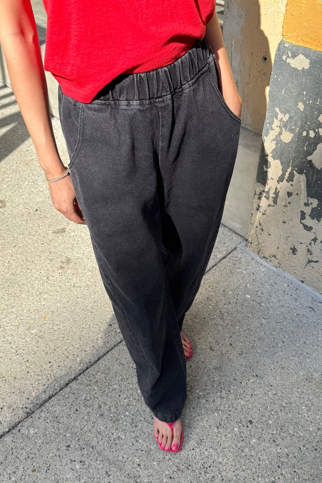 A person wearing a red shirt and Le Bon Shoppe - Long Arc Pant / Black Denim stands barefoot on a sidewalk with hands in pockets, while the ground and a textured wall appear in the background.