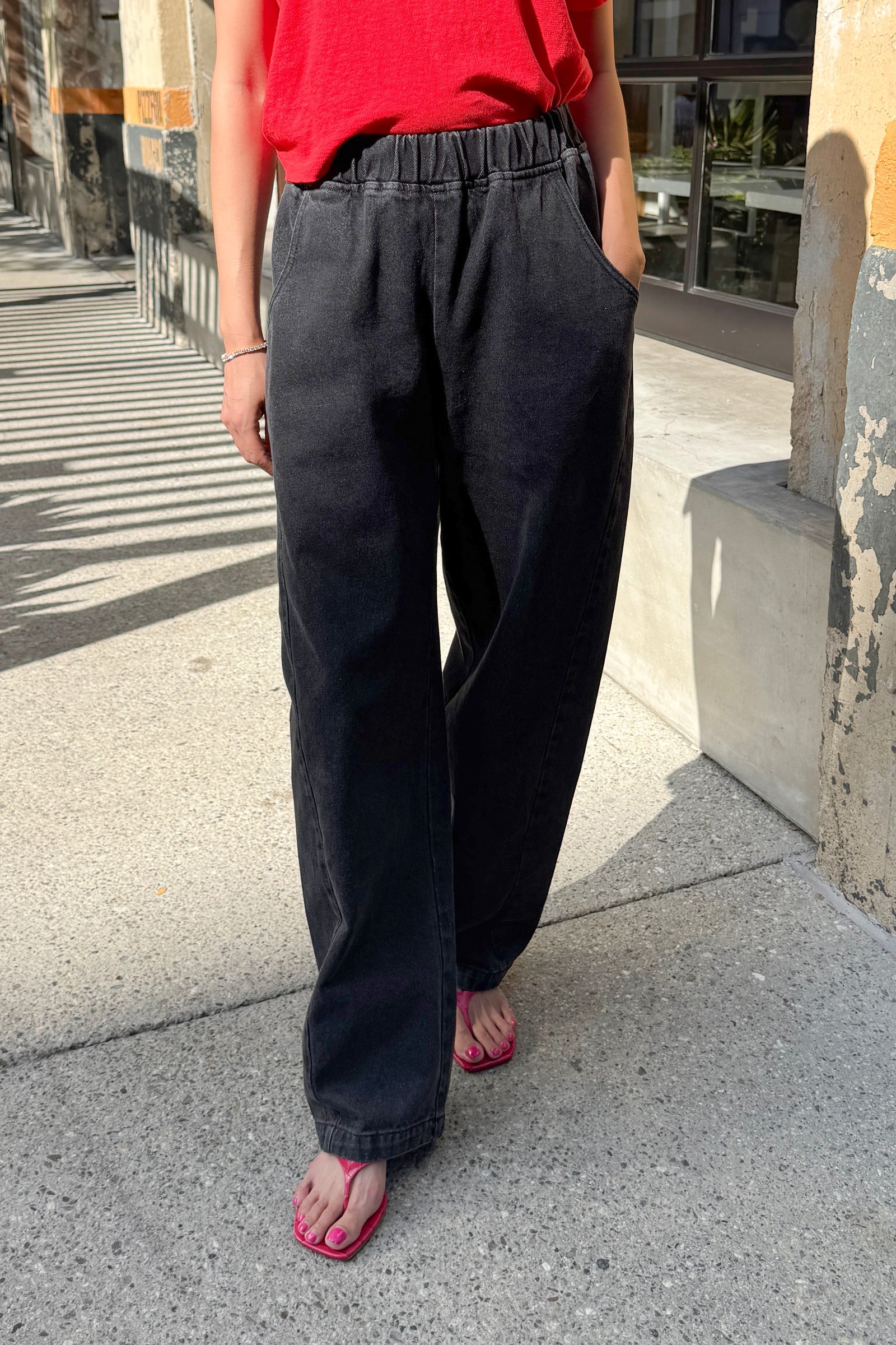 A person is seen standing on a sunny sidewalk with one hand in their pocket, wearing Le Bon Shoppe’s Long Arc Pant in black denim. Only their lower body is visible.