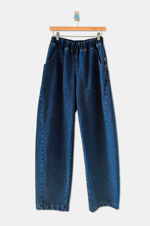 Le Bon Shoppe - Long Arc Pant / Blue Denim by Le Bon Shoppe features a dark blue, wide-leg silhouette, crafted from heavyweight denim with an elastic waistband and oversized front pockets. Displayed on a wooden hanger against a plain light background.