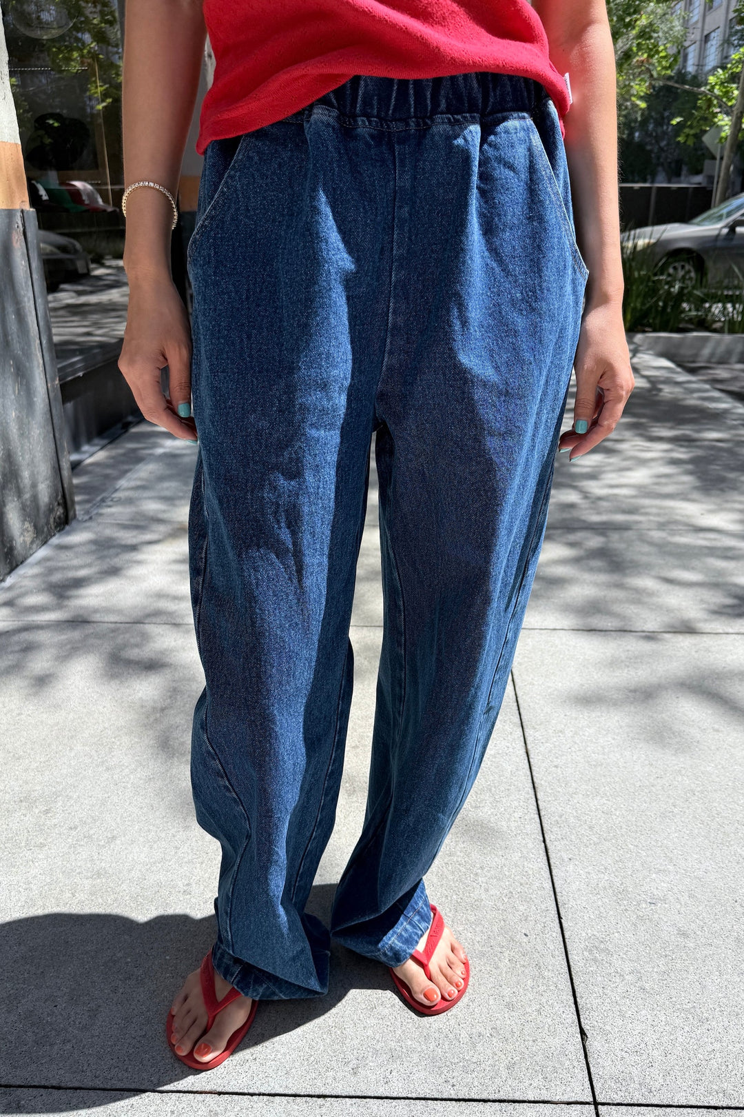 A person wearing a red shirt, Le Bon Shoppe - Long Arc Pant in Blue Denim by Le Bon Shoppe with an elastic waist, and red sandals stands on a sunny sidewalk with hands in pockets and light blue painted nails.
