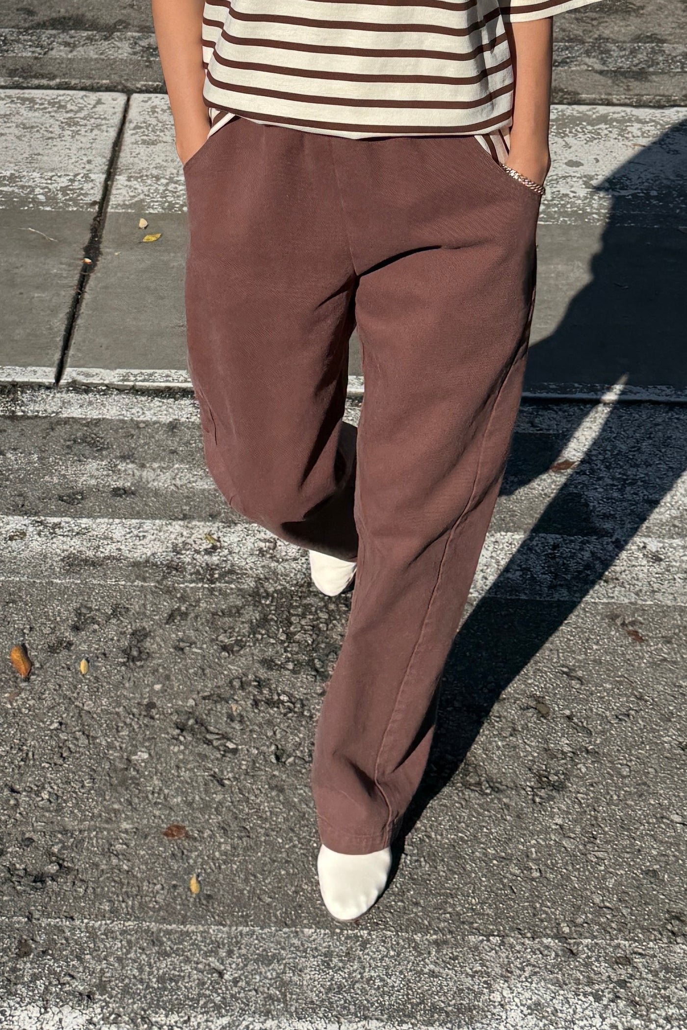 Person stands on a street crosswalk wearing Le Bon Shoppe - Long Arc Pant / Chocolate with an extended inseam, a striped shirt, and white pointed shoes, hands in pockets. Their upper face is not visible.