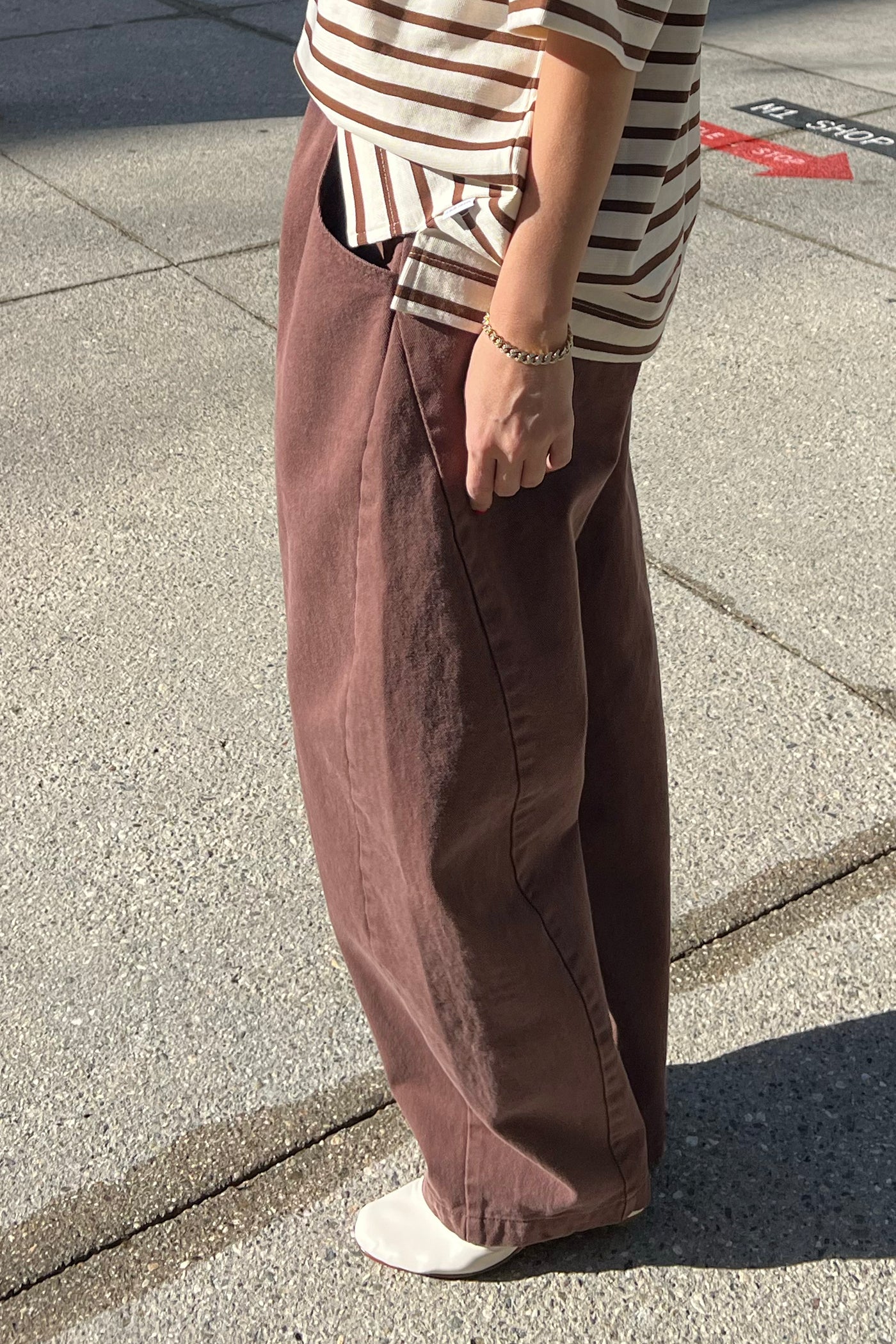 A person stands on a sunlit sidewalk wearing Le Bon Shoppe - Long Arc Pant / Chocolate, a striped beige and brown top, white shoes, and a chain bracelet. Their face is not visible.