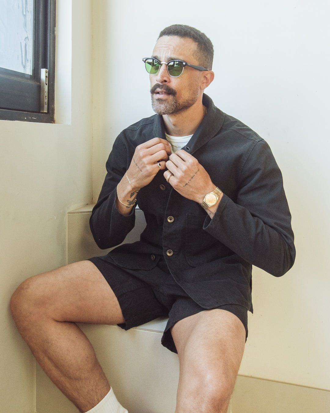 A man with a mustache and sunglasses wears an Imperfects - Shepherds Shirt Craft Canvas / Obsidian and black shorts while sitting indoors.