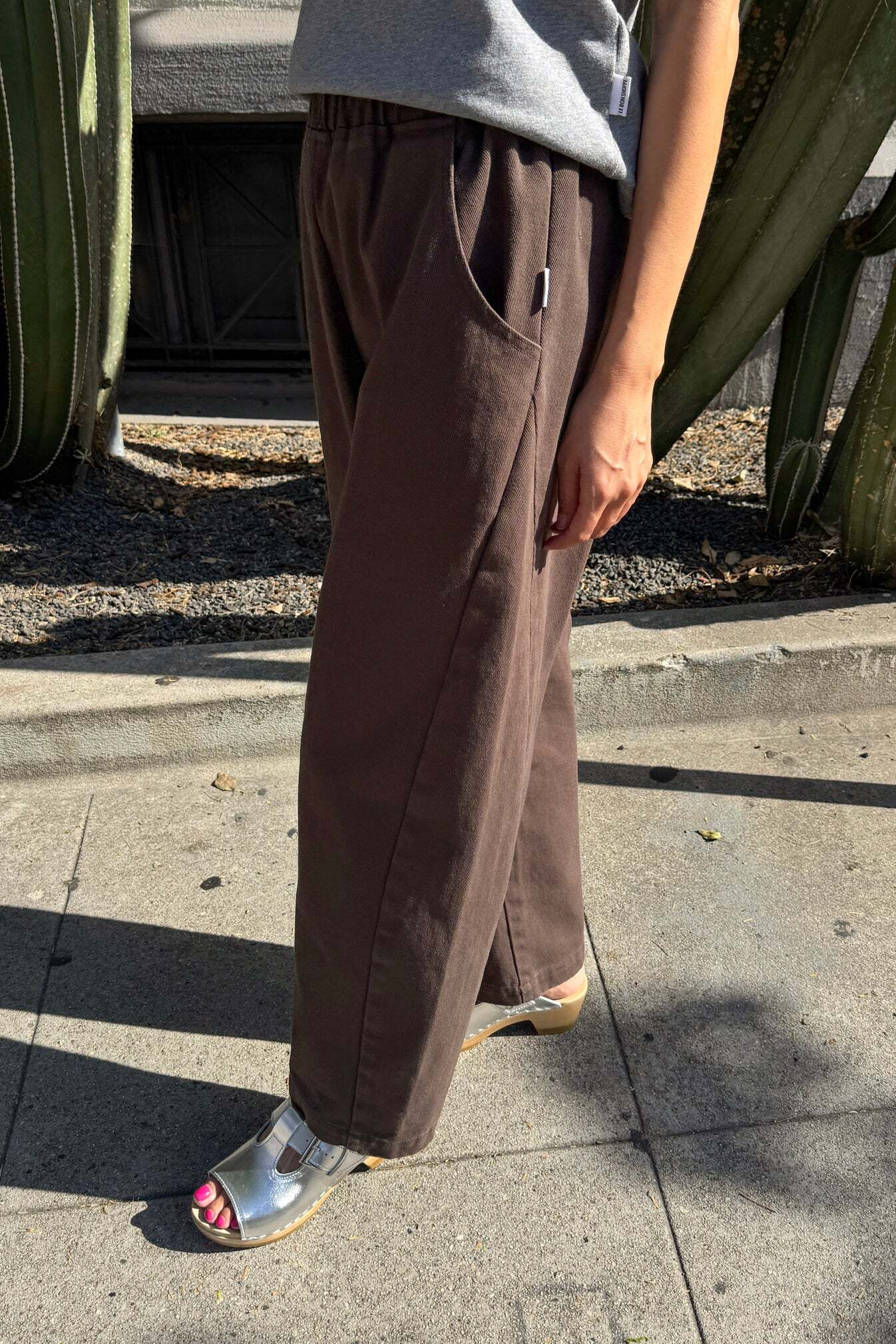 A person wearing Le Bon Shoppe - Arc Pant in Umber stands on a sunny sidewalk near large cacti, paired with a grey top and silver sandals. Only the lower half of their body is visible.