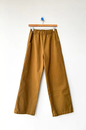 Le Bon Shoppe’s Work Pant in Dijon is a wide-leg, brown cotton canvas trouser featuring an elastic waistband and side pockets. Subtle carpenter-style details enhance its blend of practicality and style, while the pants are displayed on a wooden hanger against a plain white wall.