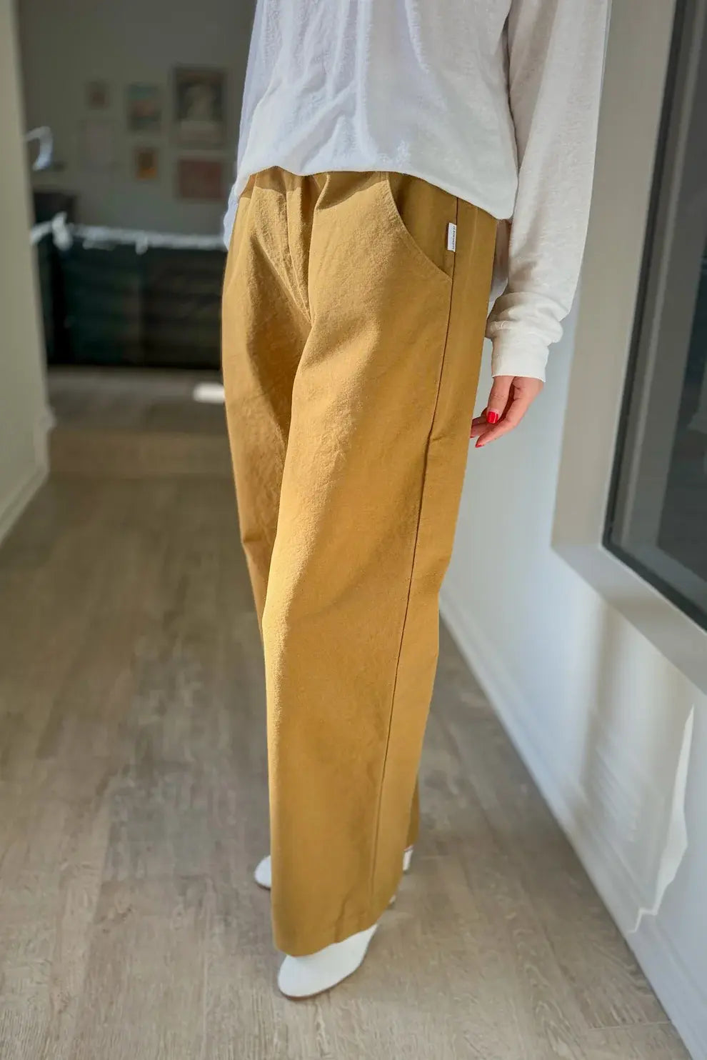 A person stands indoors with only their lower body visible, wearing Le Bon Shoppe Work Pant in Dijon—a relaxed, mustard yellow cotton canvas wide-leg carpenter style—paired with a white long-sleeve shirt and white socks, illuminated by natural light.