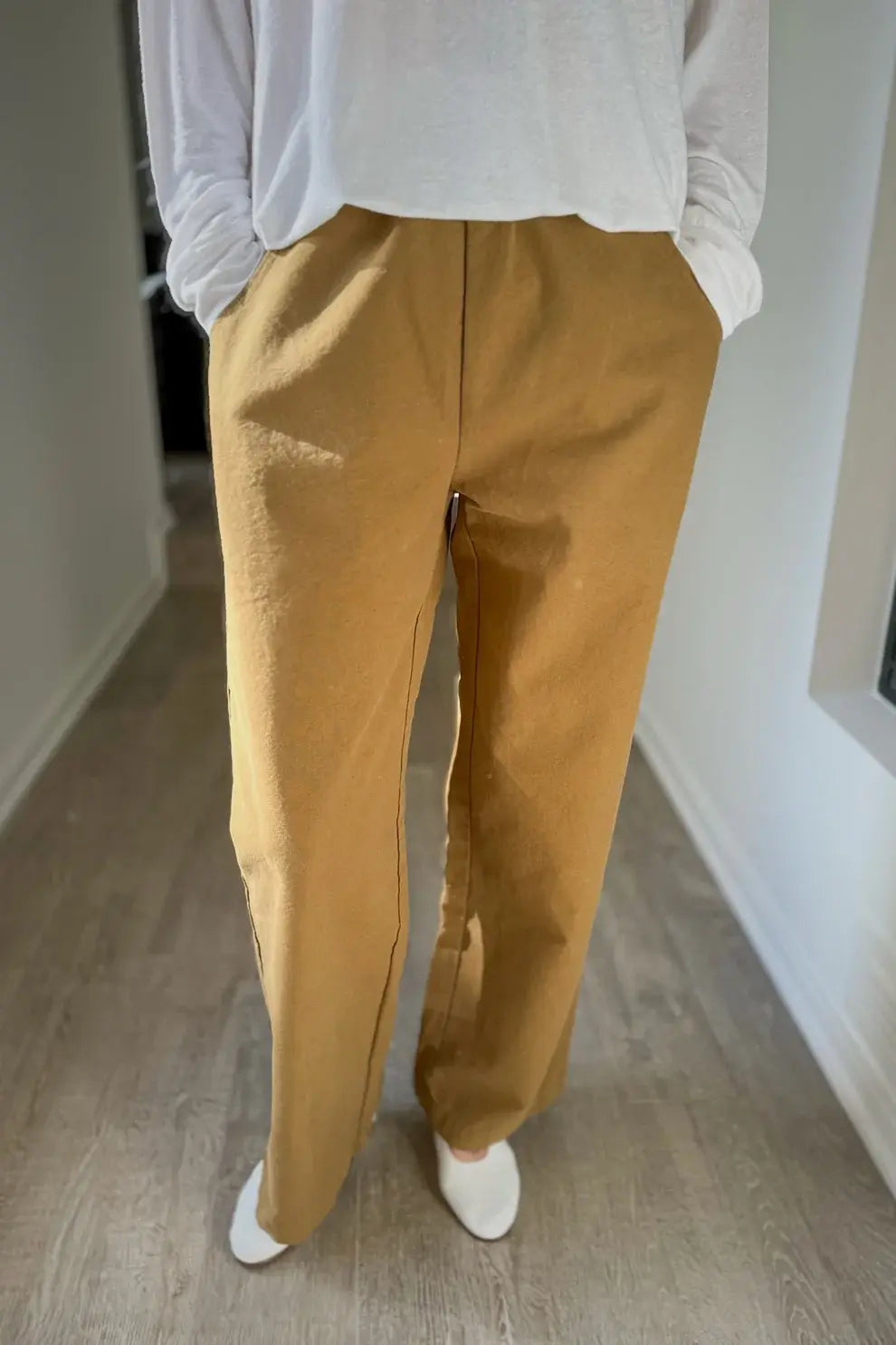 A person stands indoors on a light wood floor, hands in the pockets of their Le Bon Shoppe - Work Pant in Dijon, paired with a white long-sleeve top; their face is out of frame.