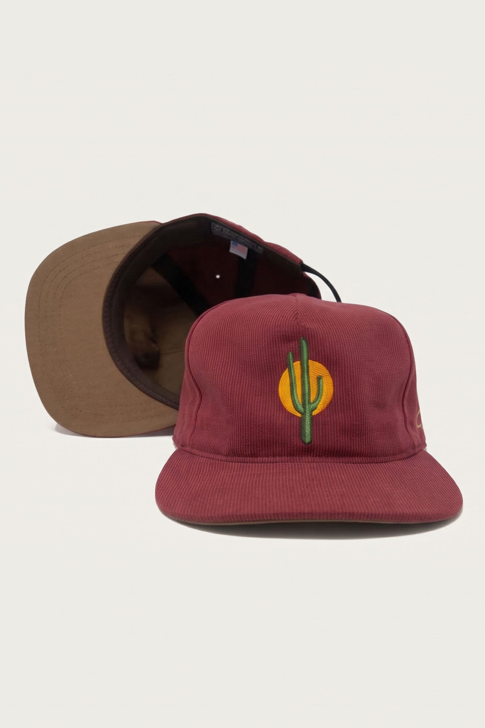 Two maroon cotton Bedford cloth The Ampal Creative - Sunset Cactus / Brick baseball caps with brown brims; the front cap features an embroidered graphic of a green cactus silhouetted against a yellow sun.