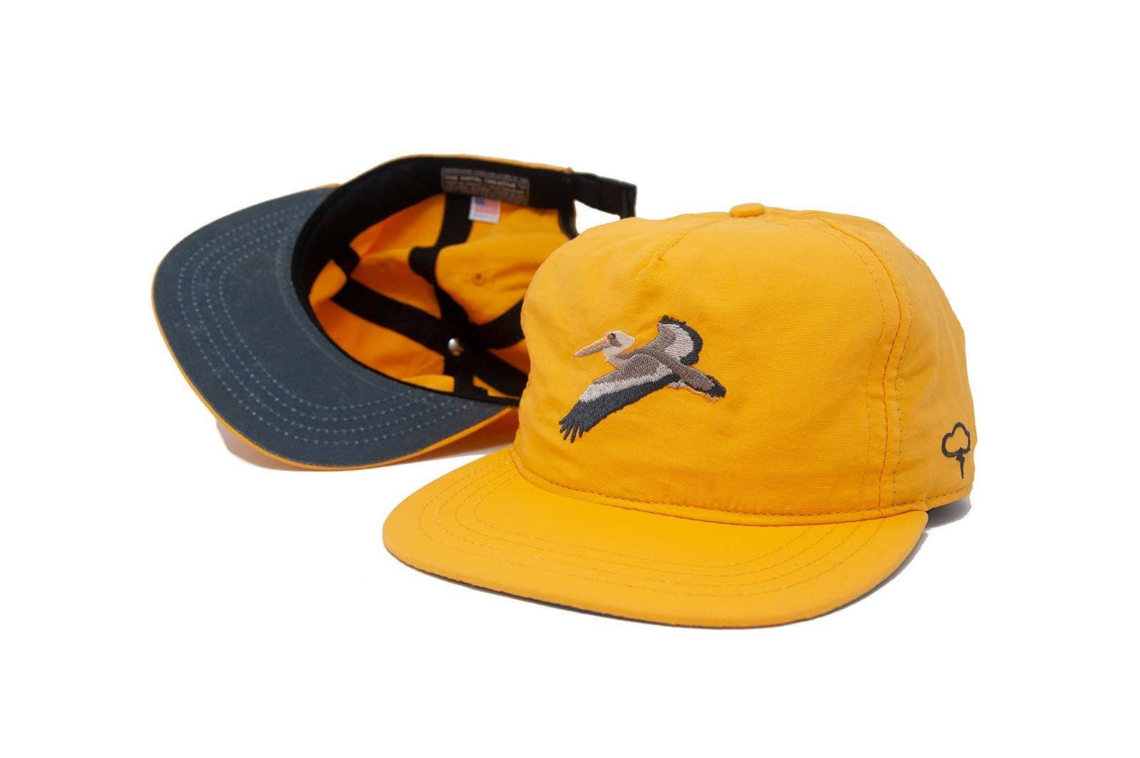 Two The Ampal Creative Glider Strapback hats in gold—one front view, one inside. Lightweight nylon with a flying bird embroidered on the front, a small tree logo at the side, and a dark green underside on the brim.