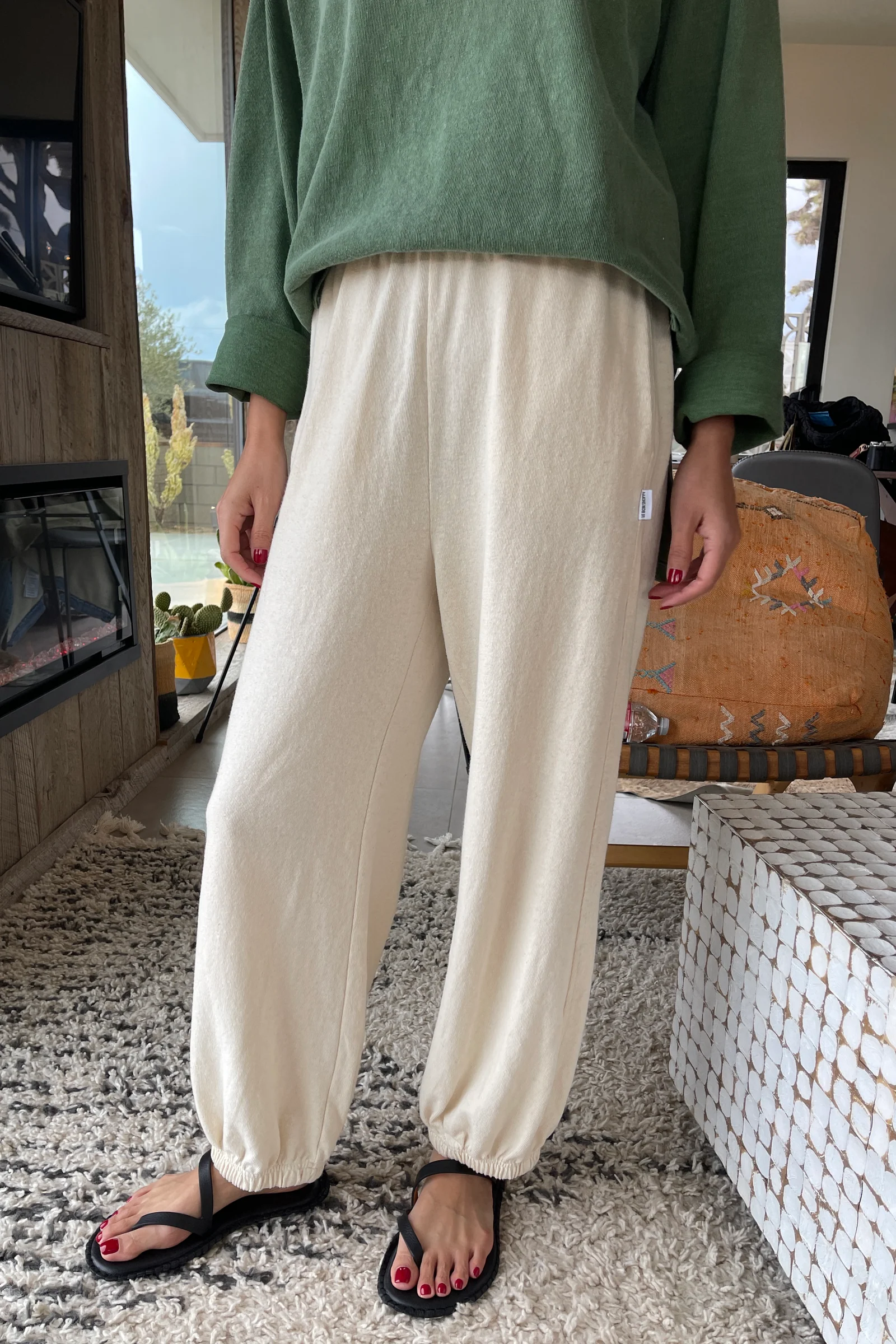 Only the lower body is visible as a person stands indoors on a textured rug, wearing Le Bon Shoppe - Balloon Pant in Natural and black sandals, with red-painted toenails.