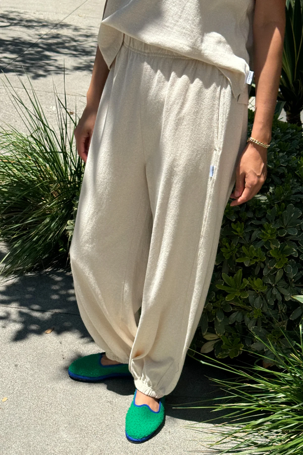 Only the lower half of a person is visible as they stand outdoors on a sidewalk by greenery, wearing Le Bon Shoppe - Balloon Pant / Natural and a matching beige top, paired with bright green slippers featuring blue trim.