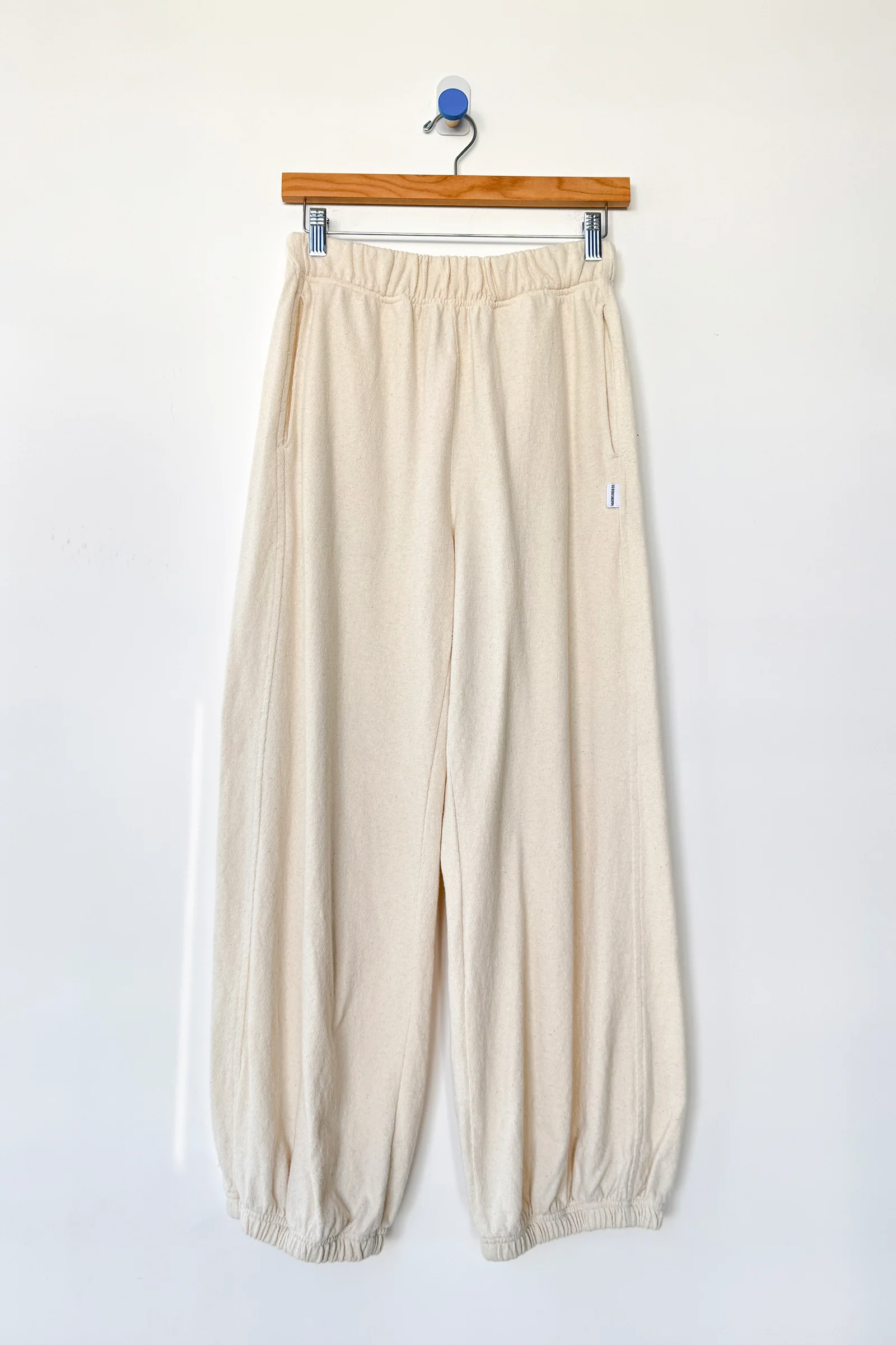 Le Bon Shoppe - Balloon Pant / Natural: Off-white, loose-fitting raw cotton pants with an elastic waistband and cuffs by Le Bon Shoppe, shown on a wooden hanger against a plain white wall.