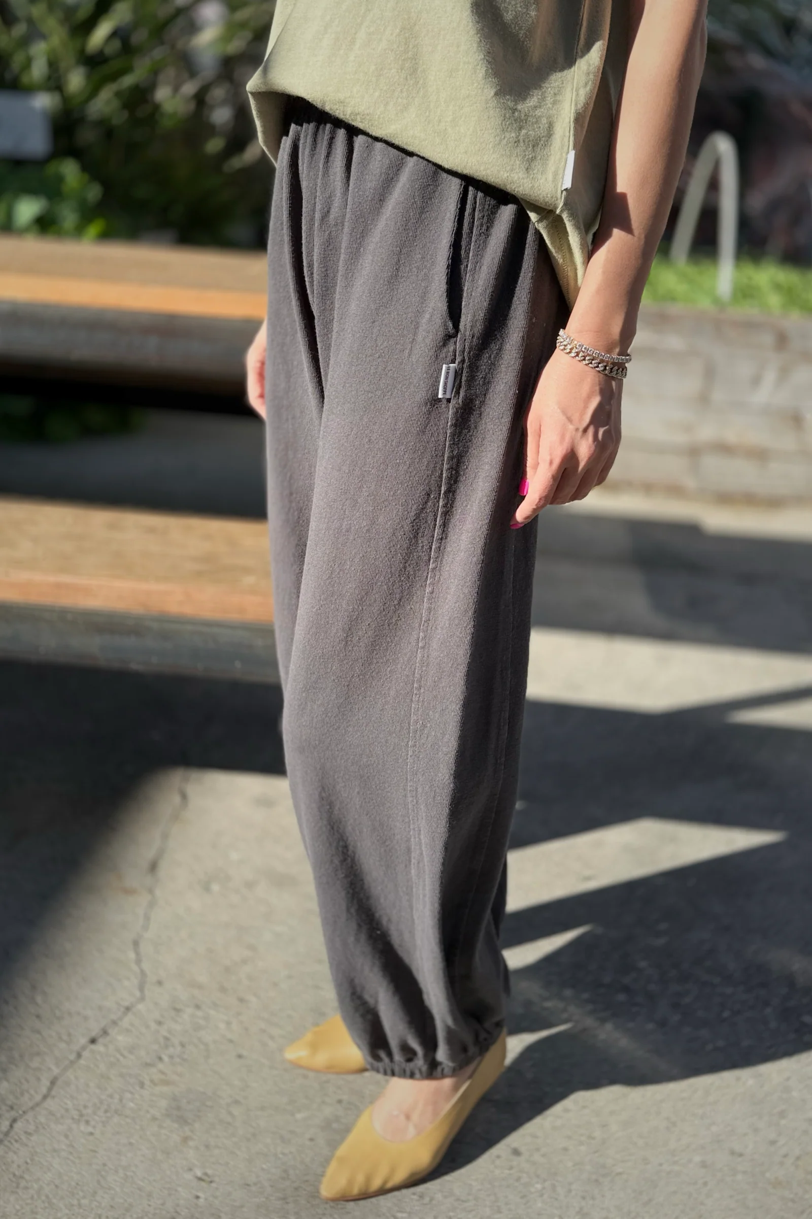 A person stands outdoors in Le Bon Shoppe's Balloon Pant in Dark Slate, paired with a light green top, yellow pointed shoes, and a bracelet, as sunlight casts soft shadows on the ground.
