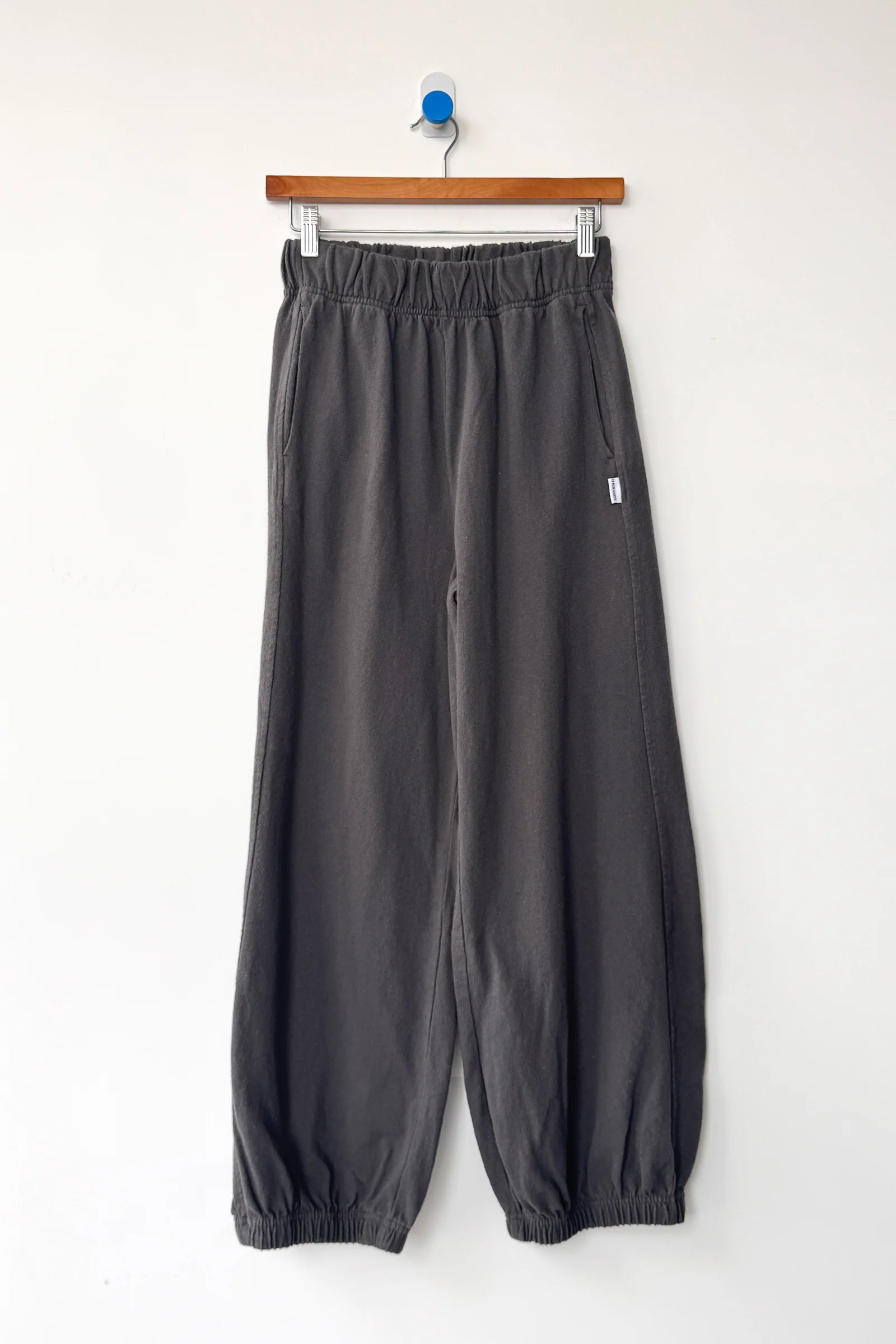 The Le Bon Shoppe Balloon Pant in Dark Slate, made from heavy cotton with an elastic waistband and cuffs, hangs from a wooden hanger on a white wall.
