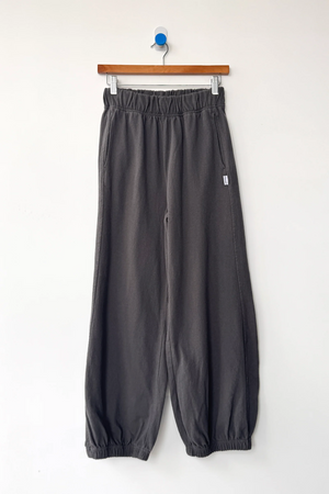 The Le Bon Shoppe Balloon Pant in Dark Slate, made from heavy cotton with an elastic waistband and cuffs, hangs from a wooden hanger on a white wall.