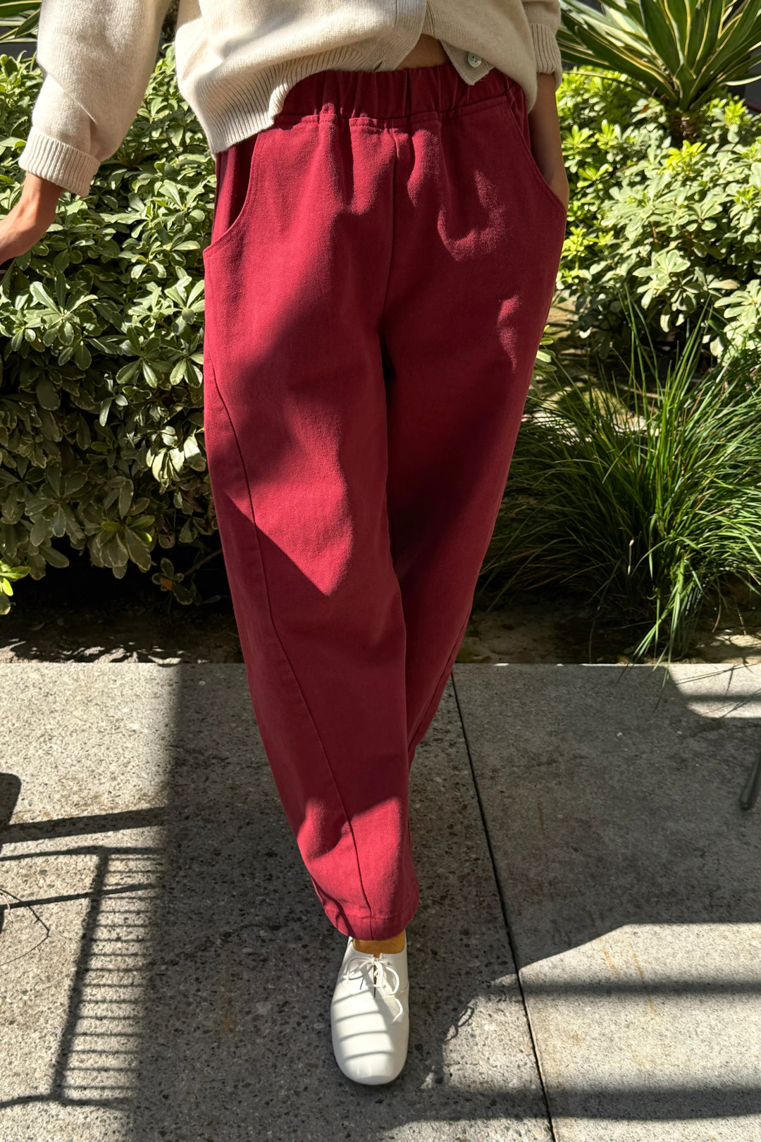 A person wearing Le Bon Shoppe Arc Pant in Oxblood, which are deep red high-waisted cropped denim pants with an elastic waistband, and white shoes, standing outdoors.
