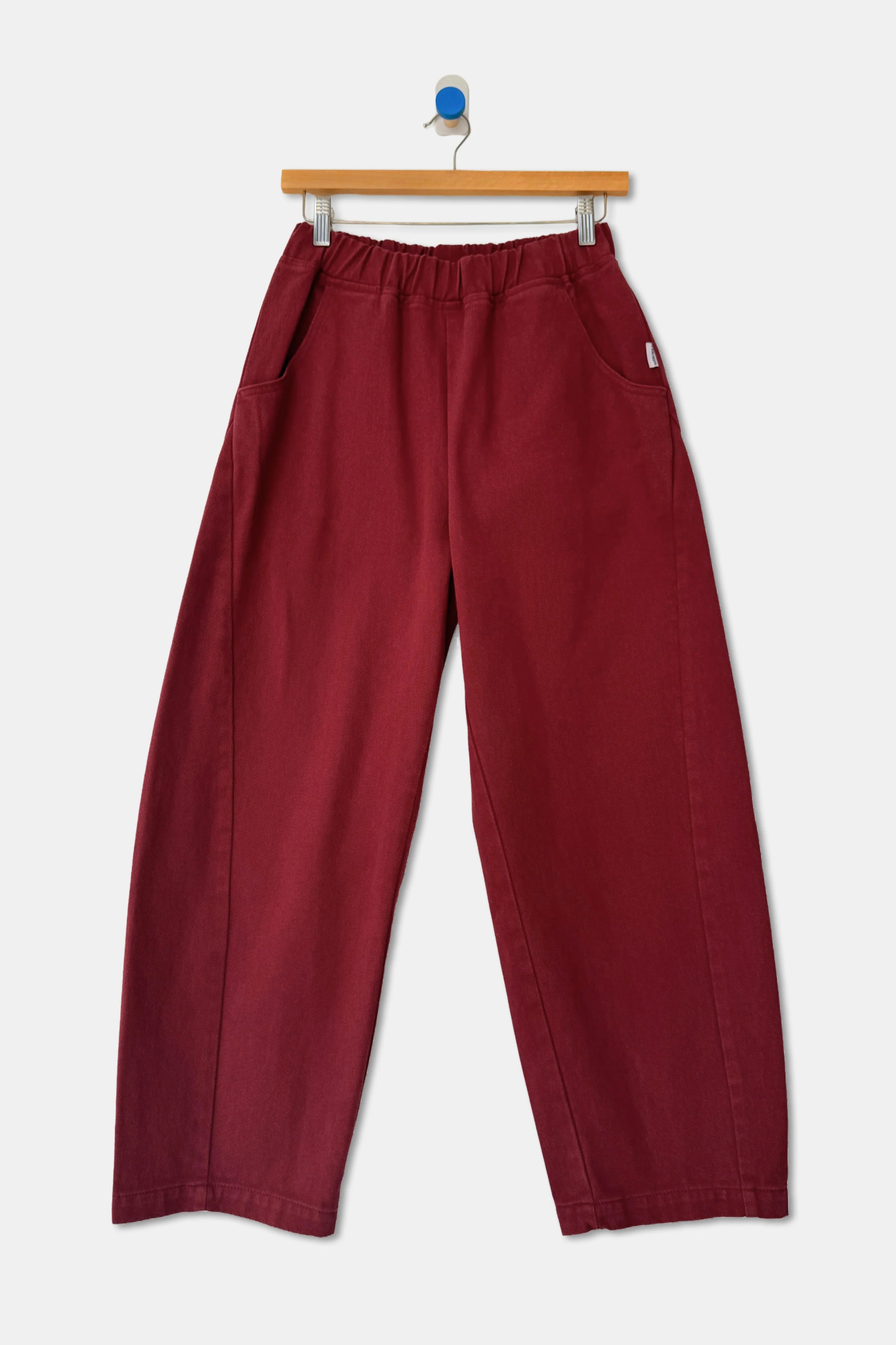 Le Bon Shoppe Arc Pant in Oxblood, featuring deep red high-waist wide-leg styling with an elastic waistband and side pockets, hanging on a wooden hanger.