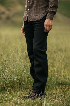 A person stands in a grassy field, wearing a brown button-up shirt and Hiroshi Kato - The Hammer Straight / Black Raw 14oz Selvedge jeans. Their dark shoes complete the look, with the blurred background highlighting their lower body and legs.