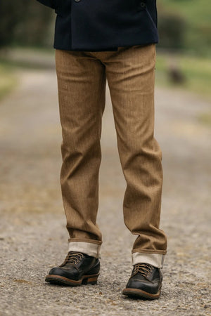 A person stands on a gravel path wearing Hiroshi Kato - The Hammer Straight / Copper Raw 10.5oz Selvedge jeans, the cuffs rolled up to reveal the Japanese selvedge denim, paired with black lace-up shoes featuring brown soles. Only their lower half is visible, with a backdrop of blurred greenery.