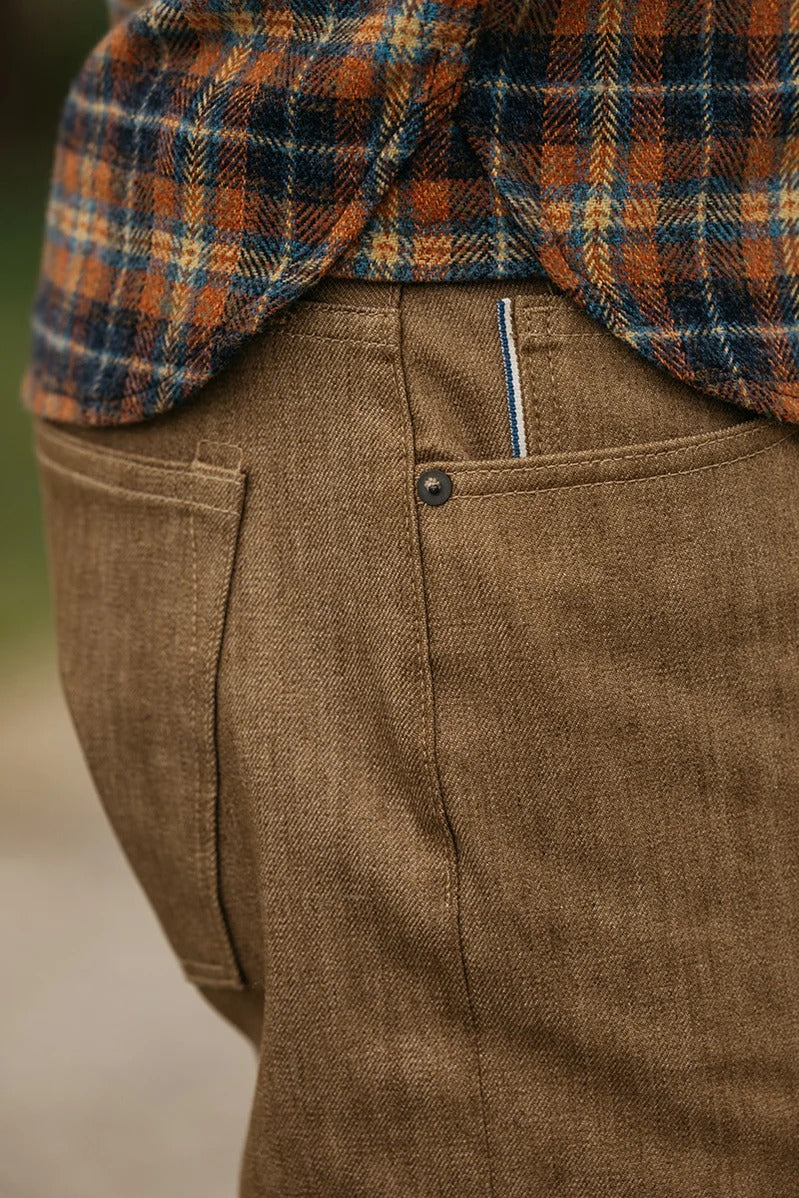 Close-up of a person wearing the Hiroshi Kato - The Hammer Straight / Copper Raw 10.5oz Selvedge denim pants, featuring back pockets, paired with a blue, orange, and white plaid shirt partially tucked in to reveal a striped tag above the pocket.