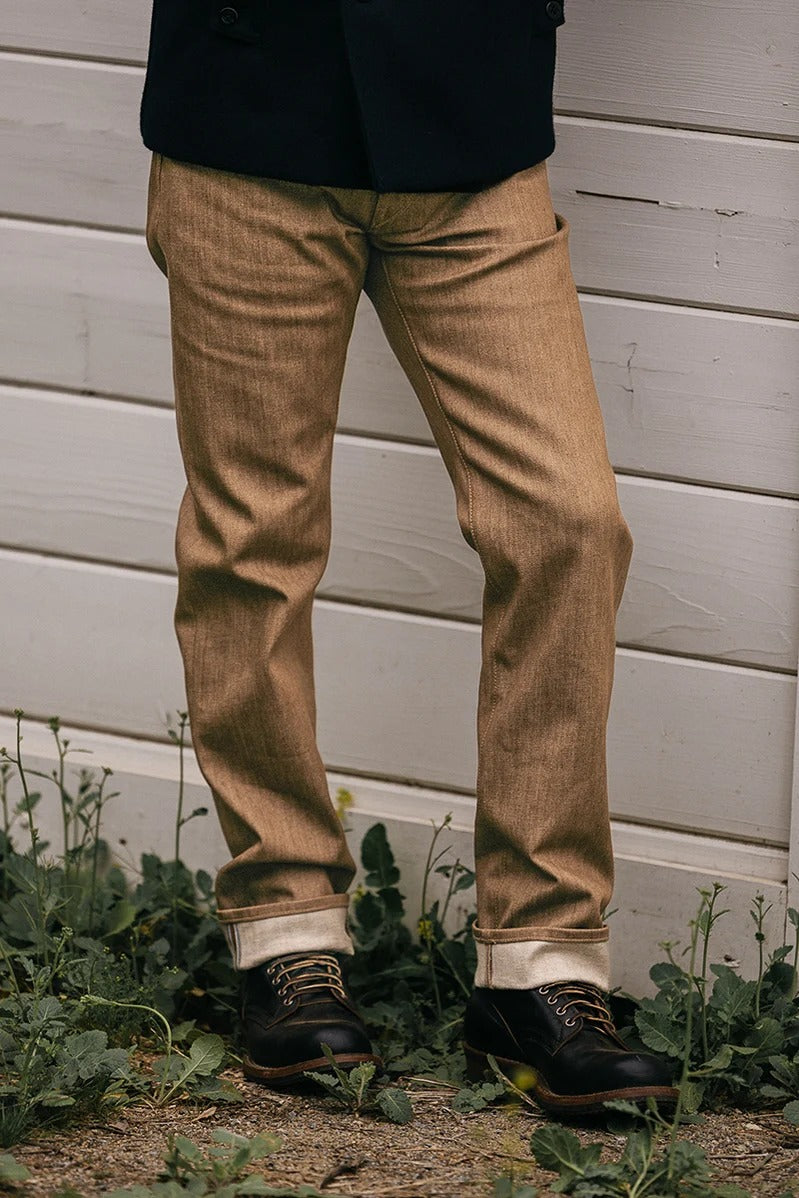 A person wearing Hiroshi Kato - The Hammer Straight / Copper Raw 10.5oz Selvedge jeans stands on a gravel surface near a white wooden wall, dressed in cuffed denim, black leather shoes, and a dark jacket, with green plants growing at their feet.